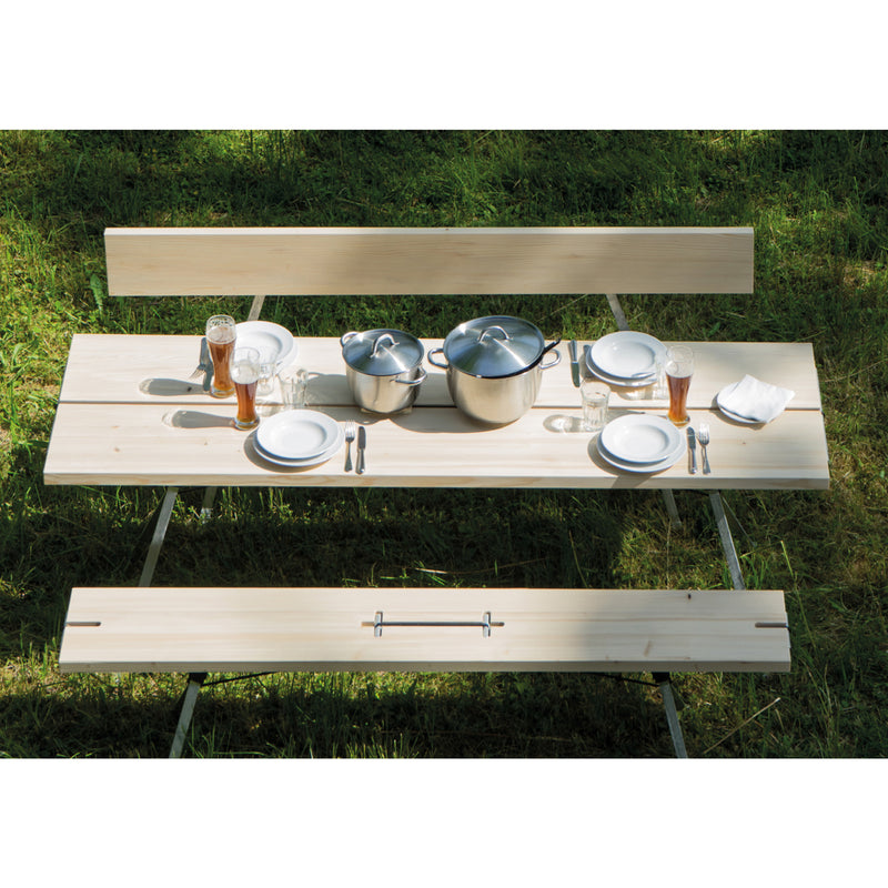 A Moormann Kampenwand table set for four includes white plates, cutlery, and glasses with a dark beverage. Two metal pots with lids sit at the center while nestled on green grass. Sunlight filters through, creating an idyllic picnic scene.