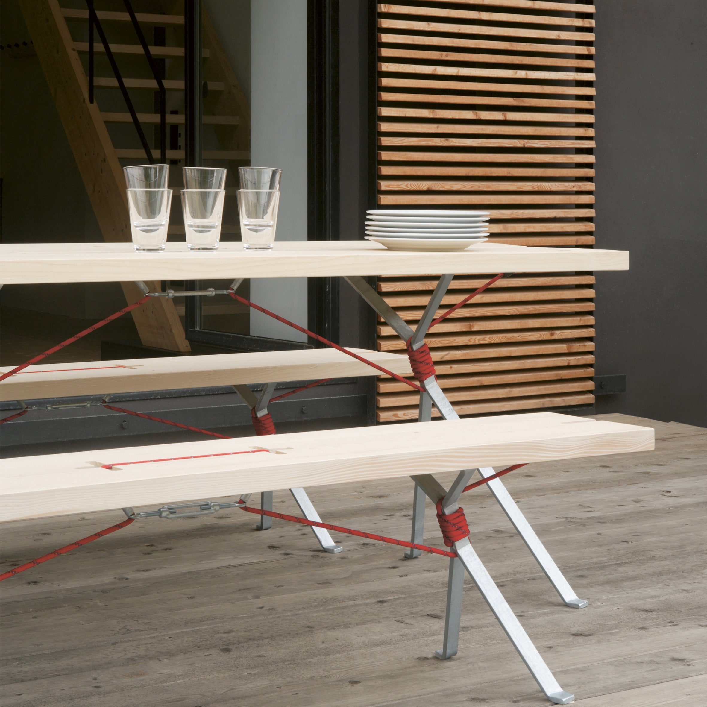 The Kampenwand table, designed by Moormann, features a light wood finish with metal legs and displays three glasses and plates. Beneath it is the Kampenwand Bench with red cords. Its situated on a wooden deck against a slatted panel backdrop.