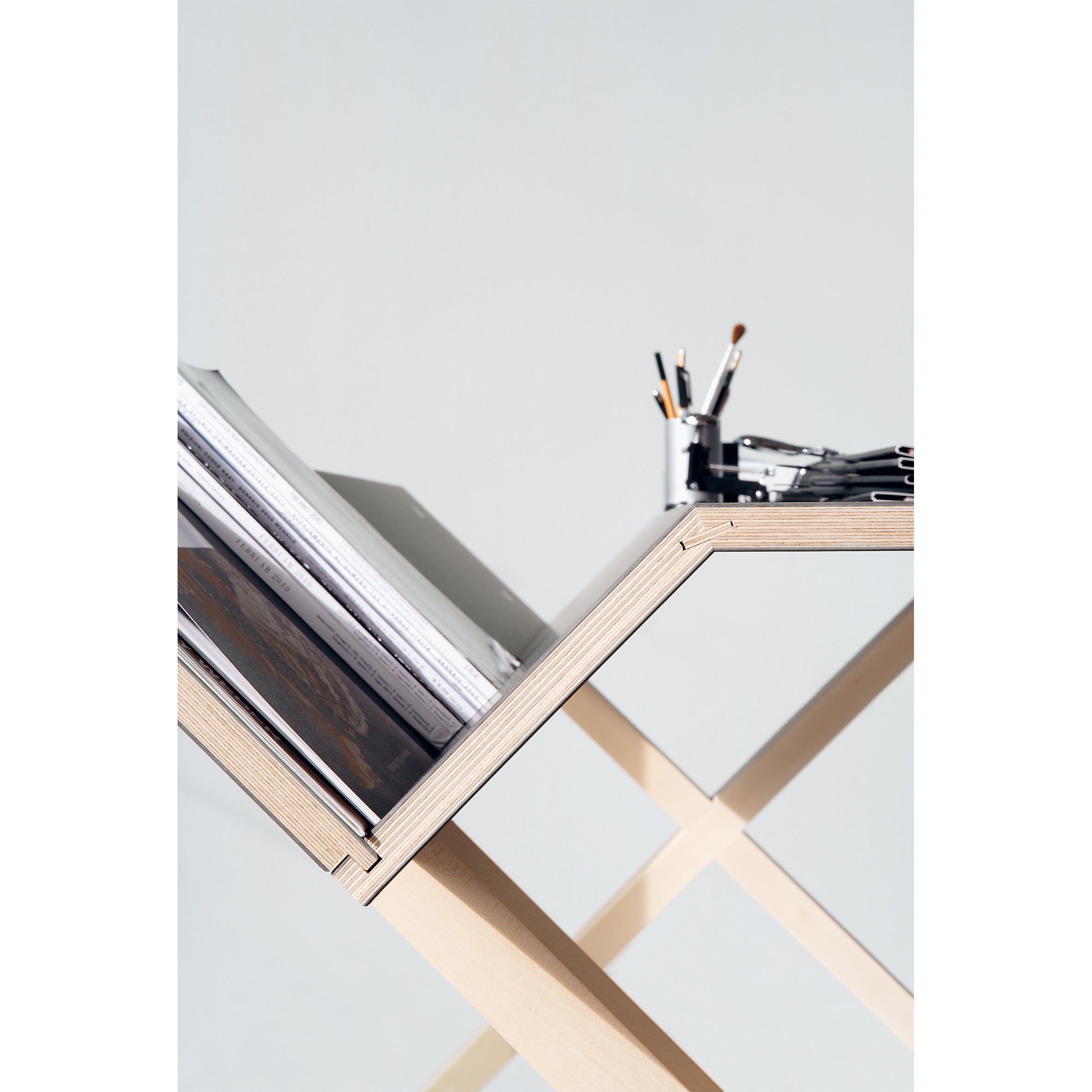 The Kant desk by Moormann features a modern wooden design with a zigzag structure, perfect for keeping workspaces tidy. Stacks of papers and a metal container with pens sit neatly on its clean surface, which is accentuated by a light gray wall.
