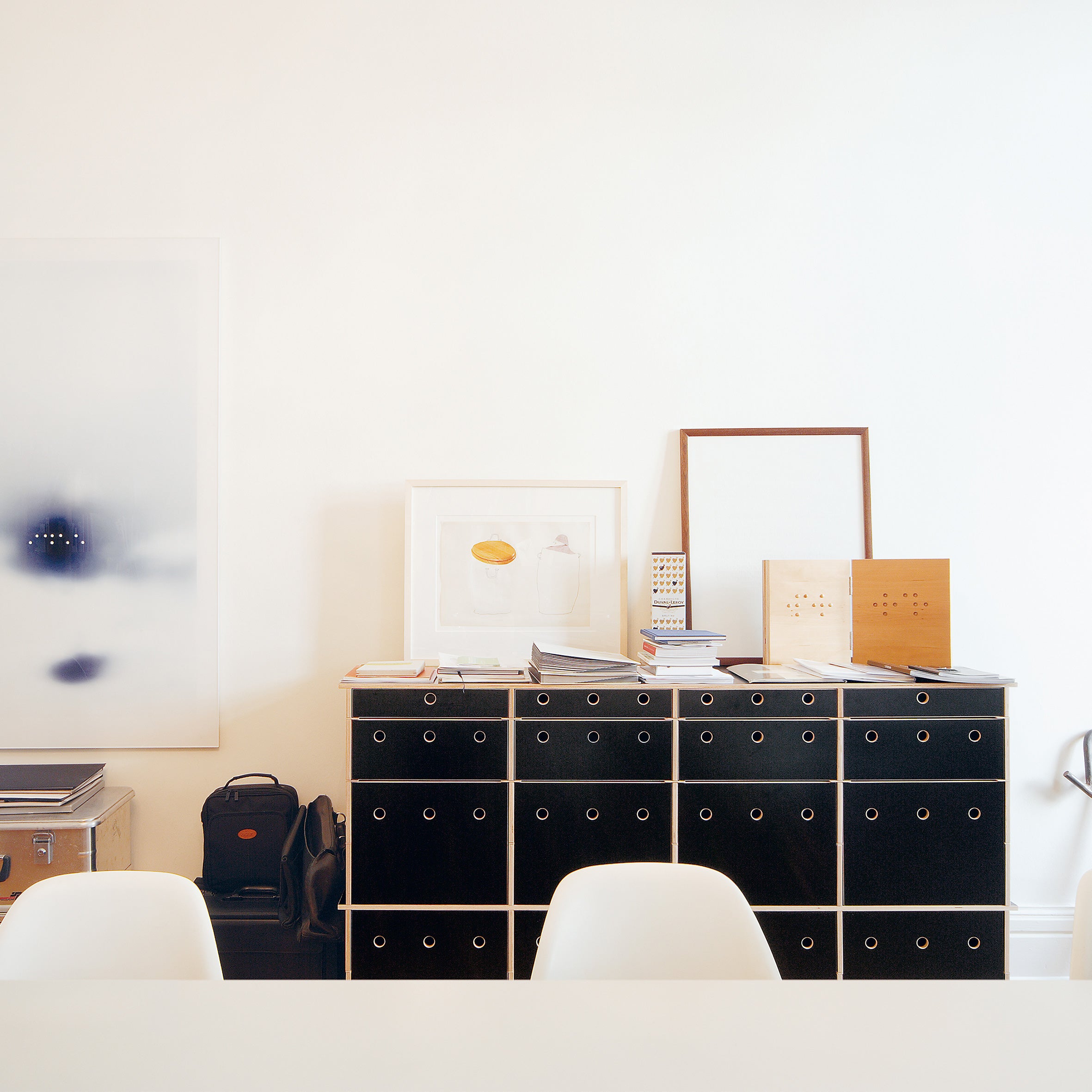 A minimalist office space features Moormanns Lader storage units with circular metal handles and customizable drawers. Artworks, books, and framed pictures rest on top. White walls and chairs create a serene atmosphere, while a backpack on the floor adds a personal touch to the modular system.