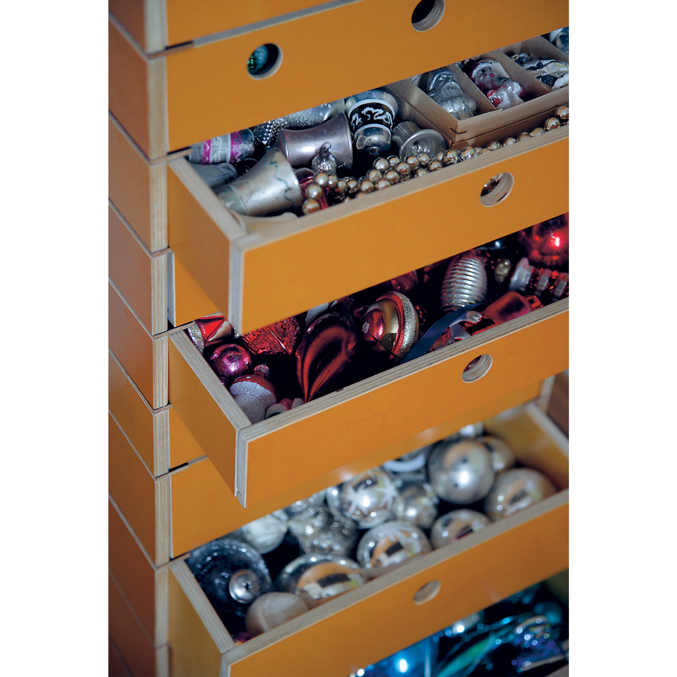 The Lader by Moormann is a modular system featuring customizable yellow birch plywood drawers with circular hole handles, filled with assorted shiny Christmas ornaments such as silver, gold, and red decorations, plus beads and ribbons.