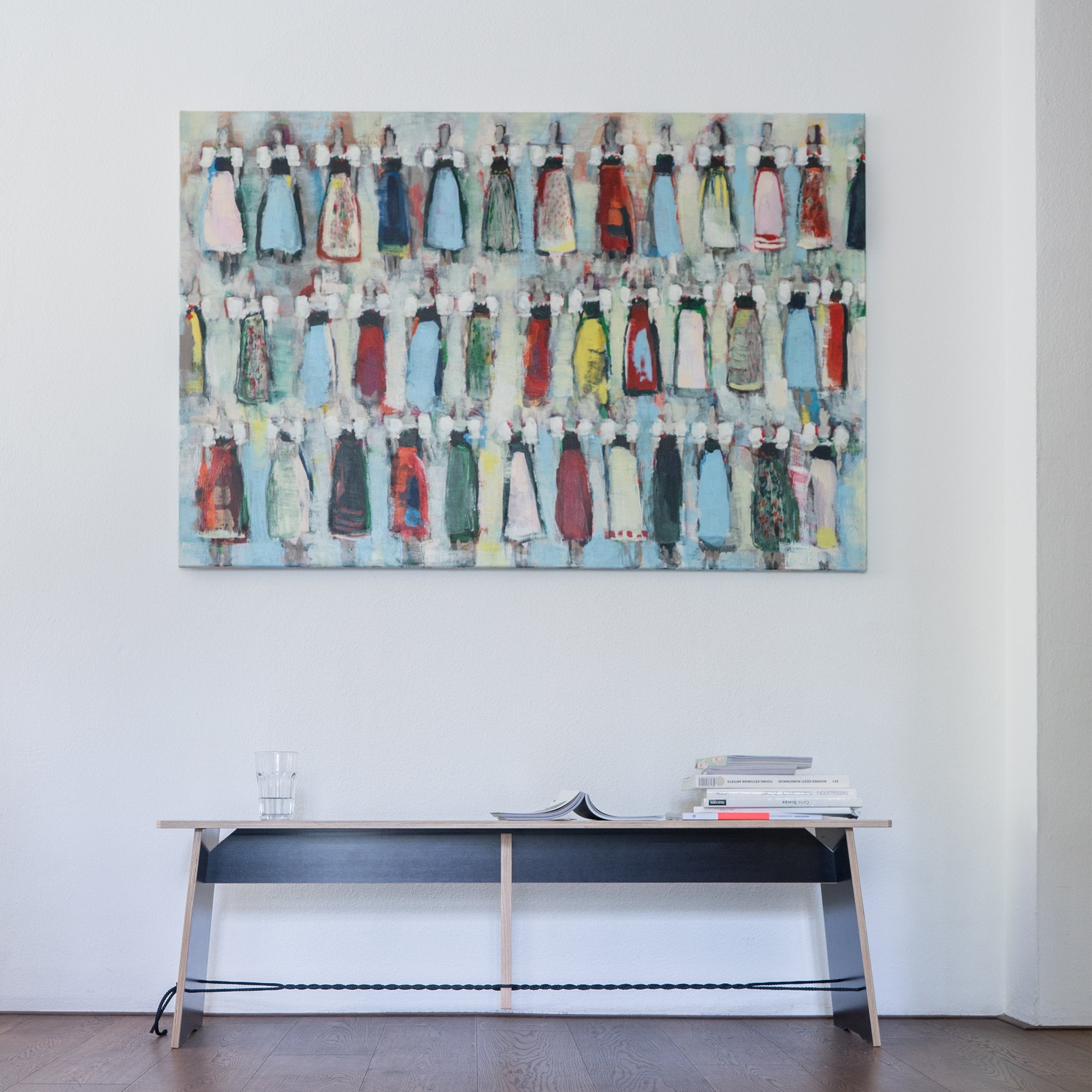 A minimalist room features a colorful abstract painting on a white wall. Below, Moormanns Langer Max bench holds a glass of water, an open book, and magazines. The wooden floor enhances the rooms clean lines and modern aesthetic.