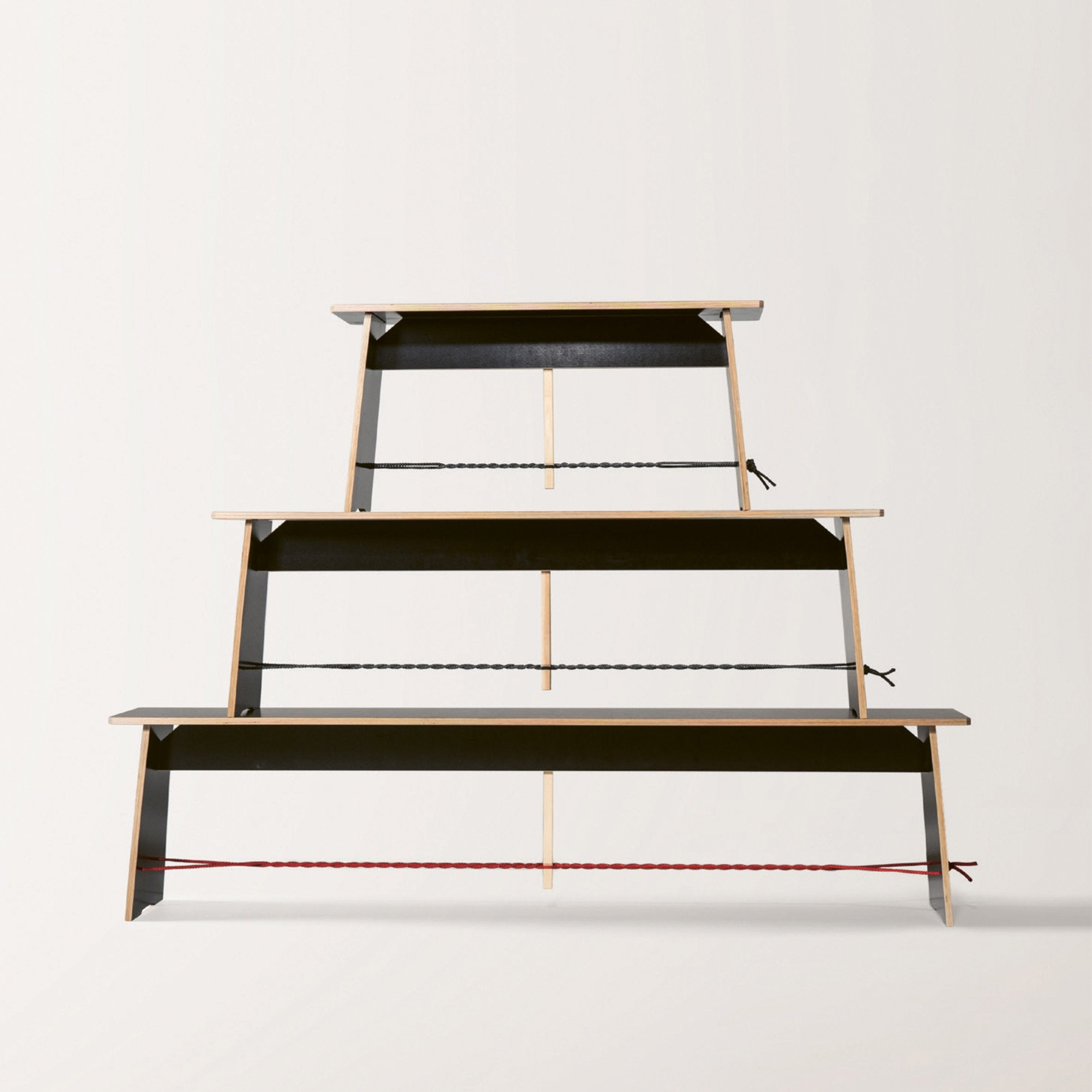 Three Langer Max benches by Moormann, crafted with designer precision, form a pyramid against a plain backdrop. Ropes support them: red at the bottom, black in the middle, and gray at the top, emphasizing their varying sizes and showcasing the artistry involved.