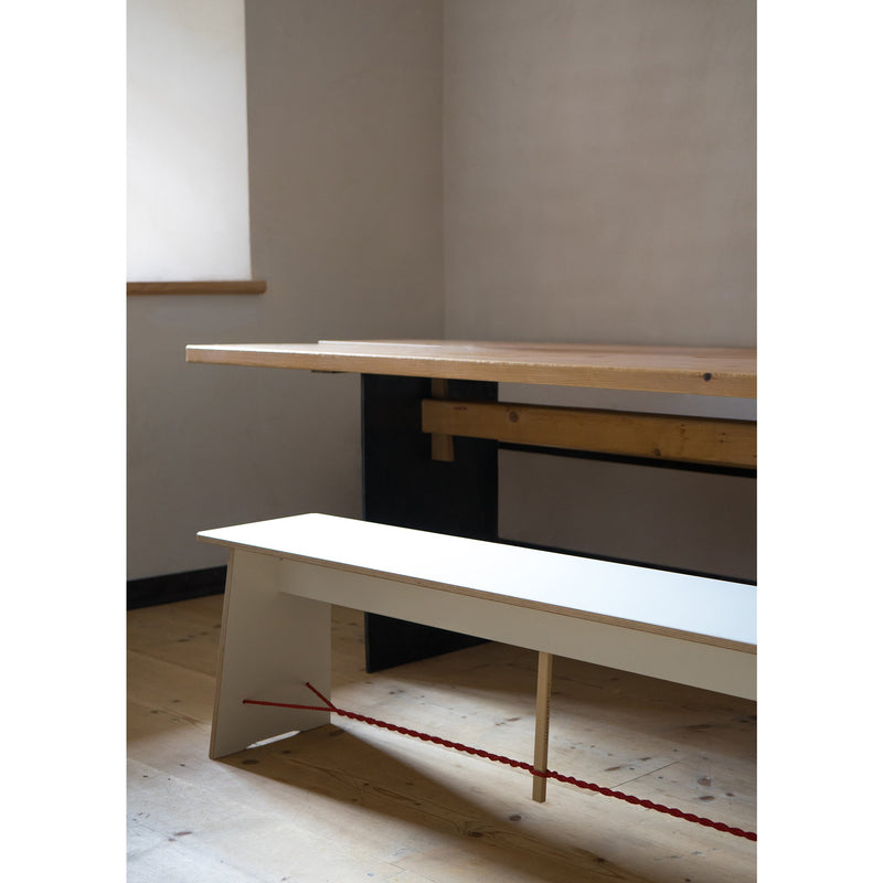 A minimalist, modern interior features Moormanns Langer Max designer furniture, including a wooden table and white bench with a red cord underneath. Light walls and wooden floors enhance the look, while soft light filters through the window for a calm atmosphere.