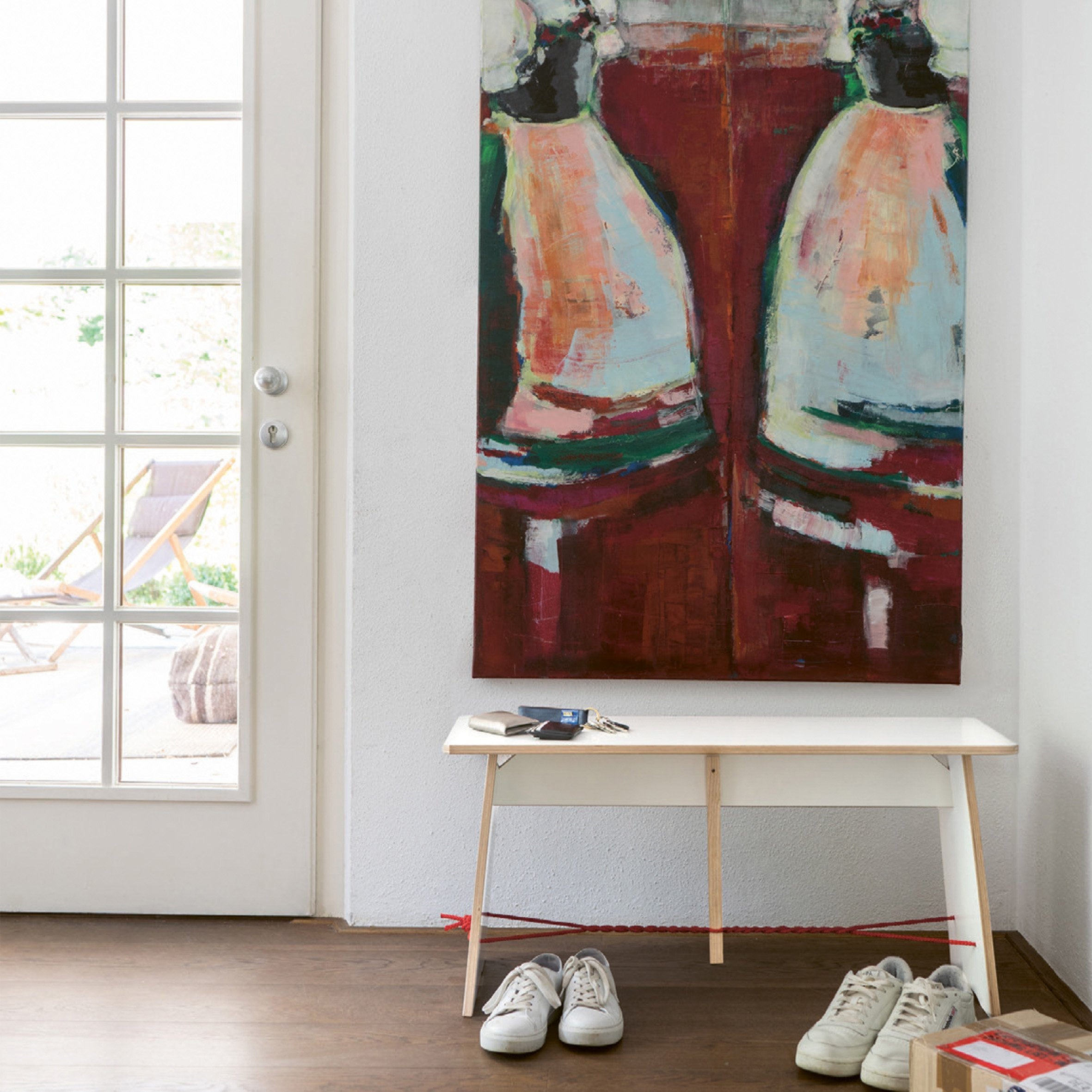 A modern painting with abstract shapes in red, white, and green hangs above a white bench with two pairs of shoes beneath it. A glass-paneled door lets natural light illuminate designer furniture, including a Langer Max lounge chair by Moormann visible outside on the rug.