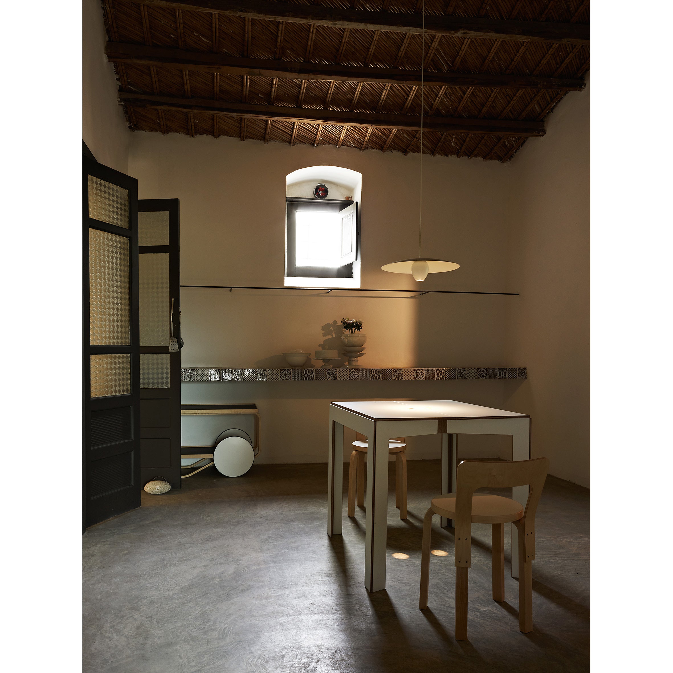 The minimalist room with a high woven ceiling showcases Moormanns Last Minute birch plywood dining table and wooden chairs beneath a modern pendant light. Natural light floods in from an open window, while simple decor like a vase and cart enhances the rustic charm of space-saving furniture.