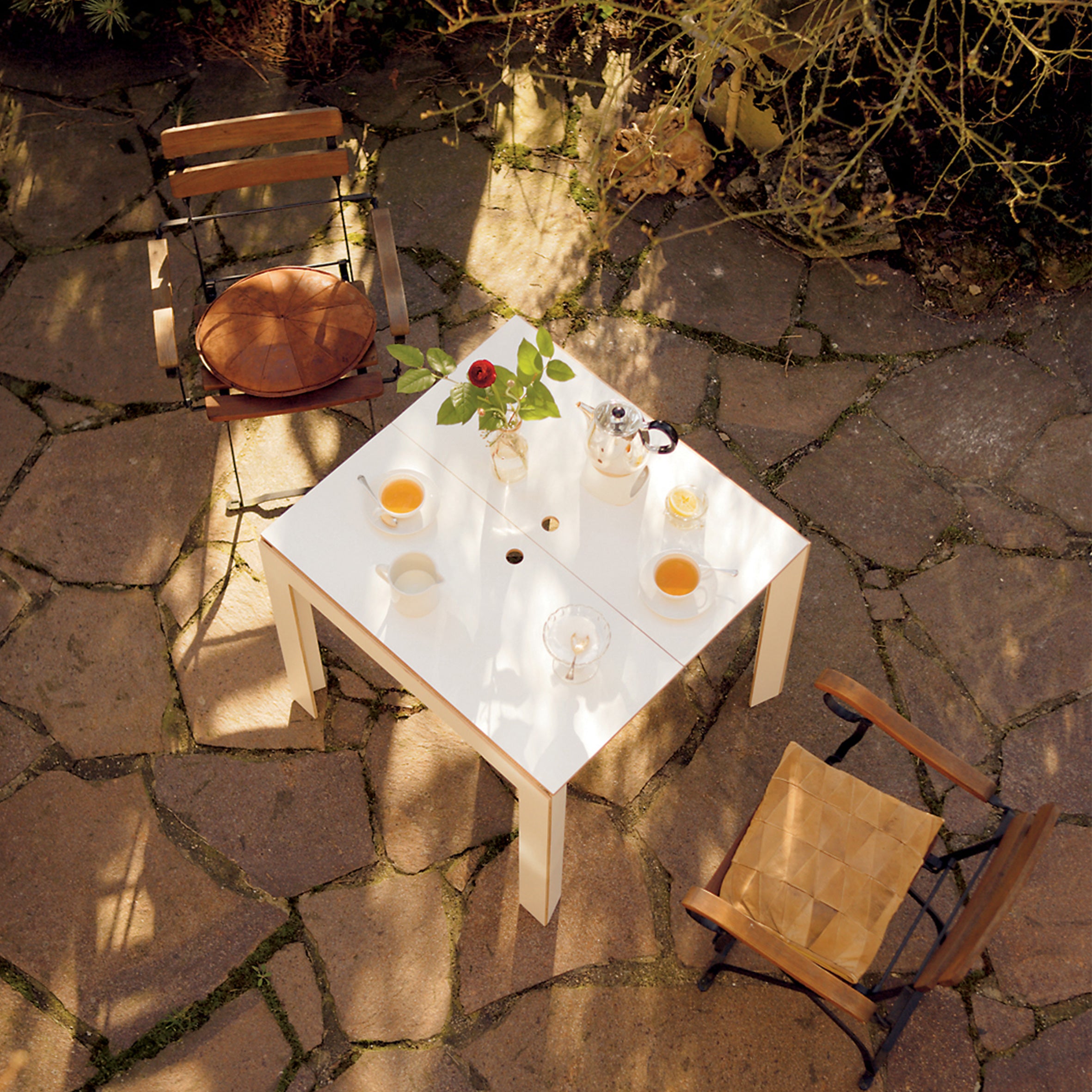 An aerial view of Moormanns Last Minute white square space-saving table features a teapot, mugs, and a vase with a red flower on a stone patio. Two cushioned chairs are positioned on either side, surrounded by plants in dappled sunlight.
