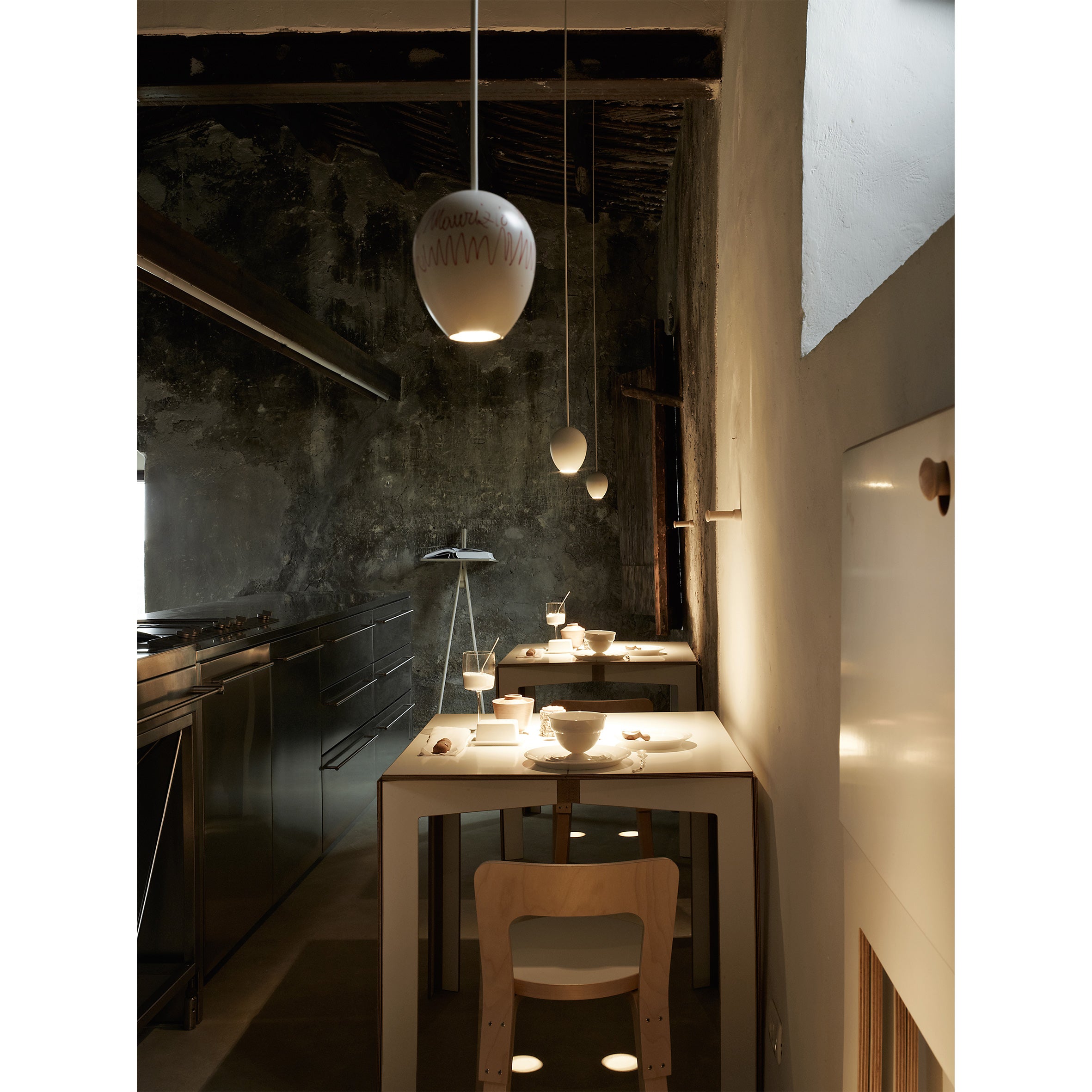 A dimly lit, narrow kitchen showcases a rustic design with a dark, textured wall and exposed wooden beams. A sleek birch plywood table by Moormanns Last Minute and wooden chairs are set for dining, illuminated by pendant lights. Stainless steel appliances line the opposite wall.