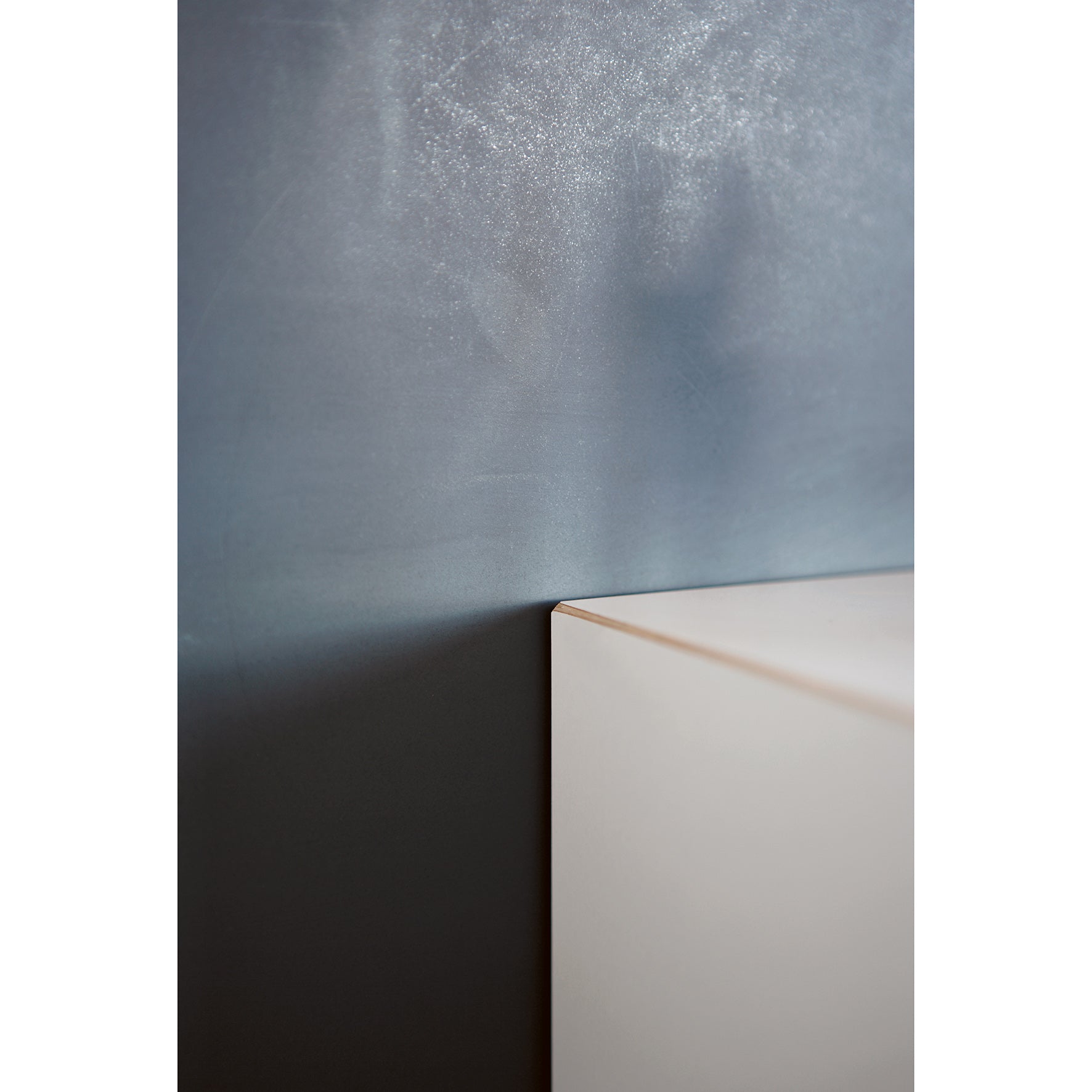 The Magnetique shelf by Moormann, a sleek, light-colored rectangle with integrated magnets, is positioned against a textured dark wall. Its sharp corner casts a faint shadow on the surface.