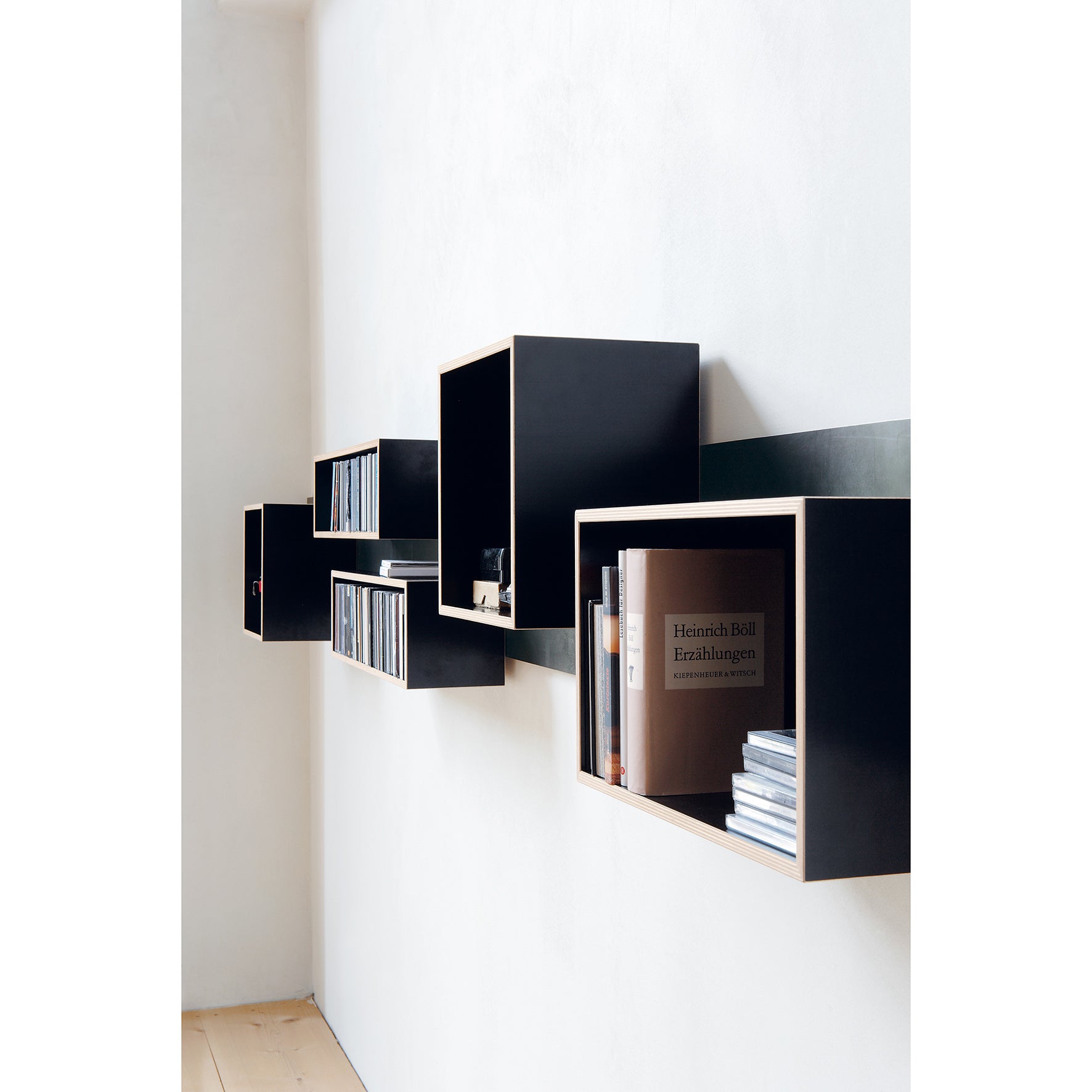 Moormanns Magnetique shelves, with a black wall mount and light wood trim, feature integrated magnets to hold books and CDs in a staggered pattern on a white wall. The natural light highlights the minimalist design and wooden floor below.