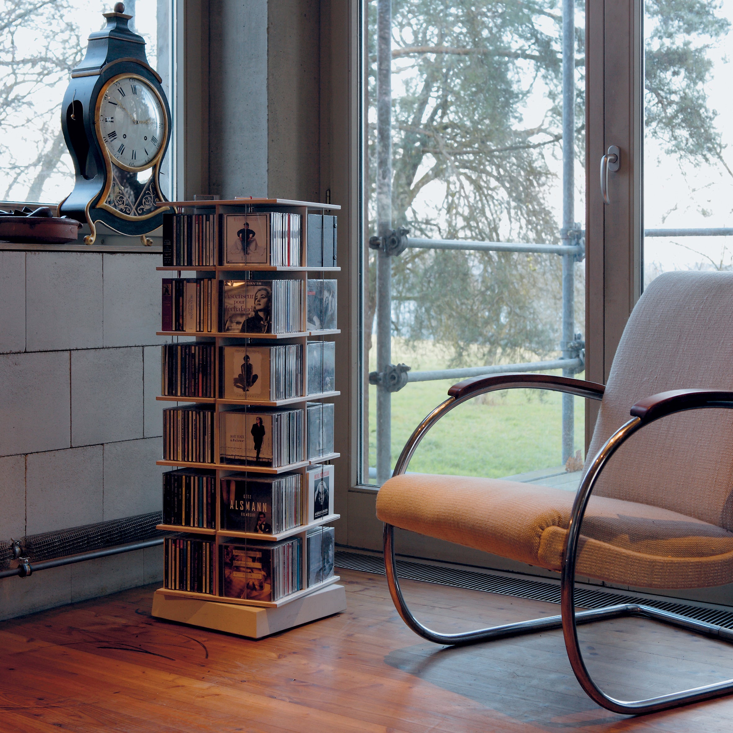 A cozy room features a chair, a vintage-style clock on a shelf, and a Moormann Musikstapler CD rack with CDs. Large windows showcase green trees, filling the room with natural light. The wooden floor adds warmth to this space-efficient design.