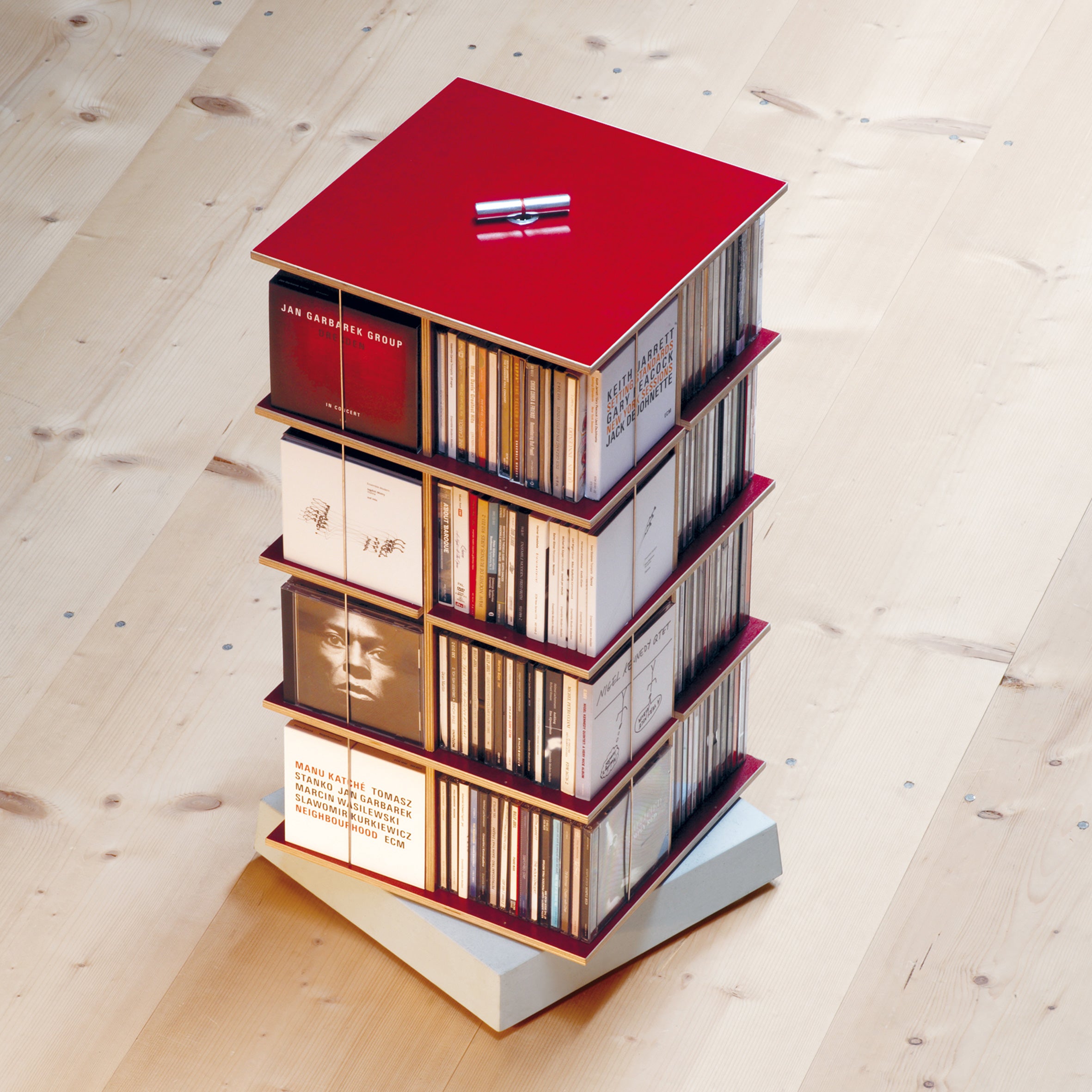 The Moormann Musikstapler, a modern space-saving CD rack with four tiers, is rotatable and red. It sits on a light wooden floor and showcases various album covers. Topped with a small metal sculpture, it elegantly holds CDs.