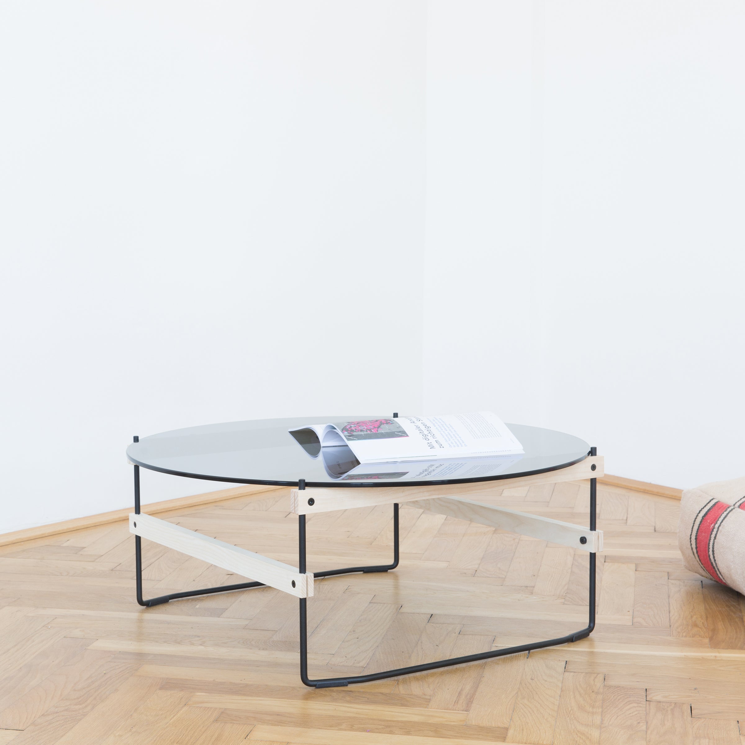 The Netto Coffee Table by Moormann stands in a corner on a wooden floor, its modern glass top with a sleek metal frame showcasing an open magazine. Made from grey tinted ESG glass, it complements the minimalist white walls. To the right, a rolled-up rug adds texture to this tasteful setup.