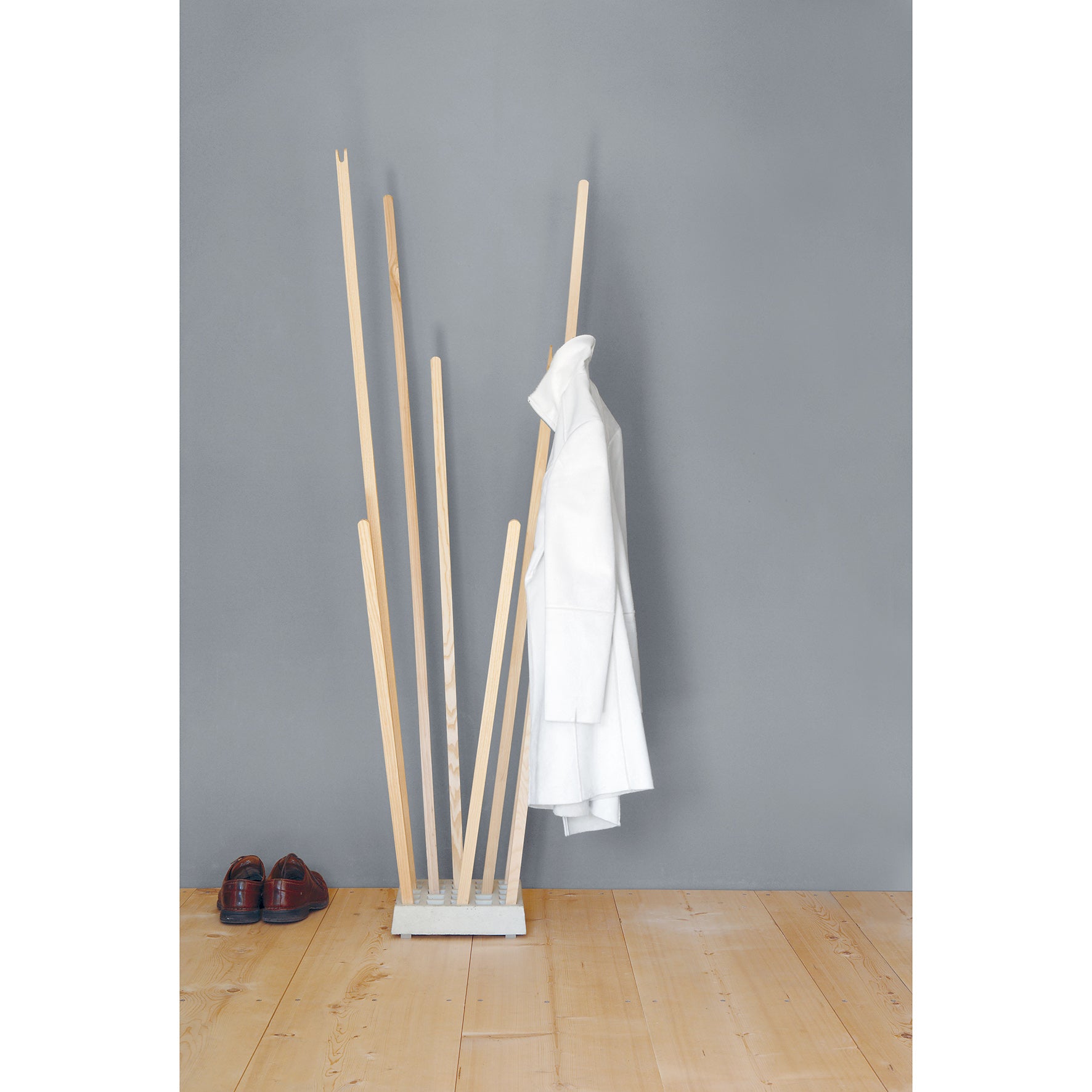 The Moormann Pin Coat, a minimalist wooden coat rack with versatile design and varying heights, stands against a gray wall and light wood floor. A white coat is draped over a peg, with dark brown shoes placed on the ground nearby.