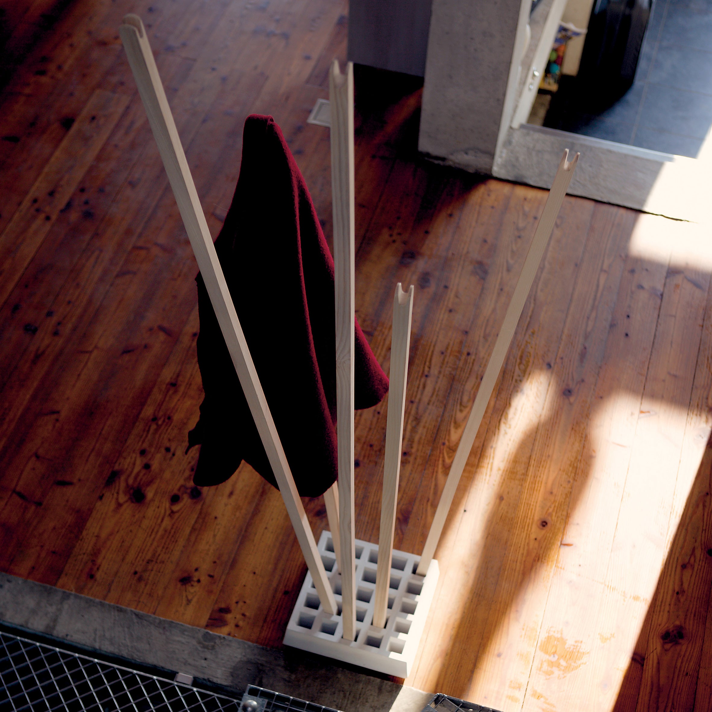 The Moormann Pin Coat, a modern wooden coat rack with tall angled pegs, supports a dark red garment on a wooden floor as sunlight streams through a nearby window.