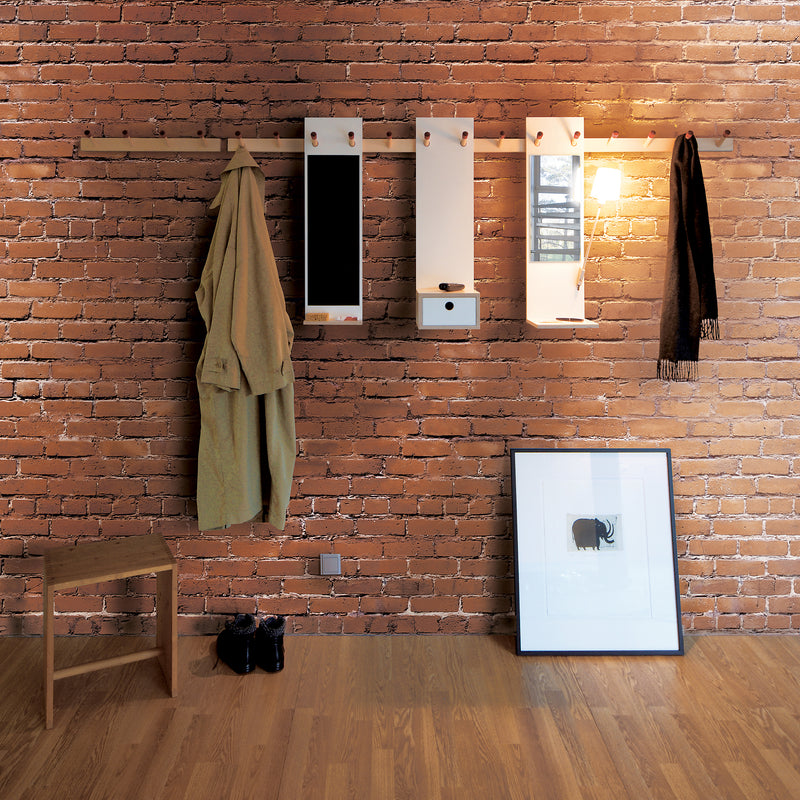 In a rustic room with a brick wall, the Moormann Rechenbeispiel hook rail holds a tan coat and scarf, with small mirrors above its flexible storage drawer. Below, black shoes are beside a wooden stool and a framed picture rests on the warm wooden floor.