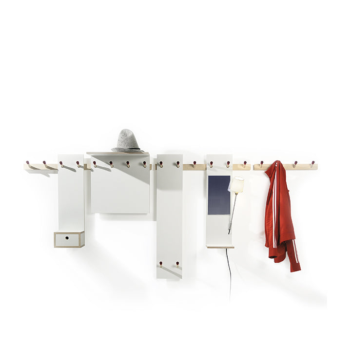 The Rechenbeispiel by Moormann is a minimalist, modular wall-mounted coat rack with multiple pegs, holding a grey hat, red jacket, and small lamp. It features flexible storage with a small compartment and vertical mirror against a plain white background.