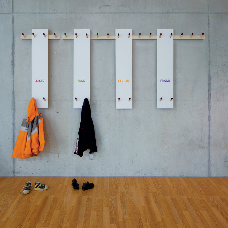 The Rechenbeispiel by Moormann is a modern coat rack with a modular system on a concrete wall, featuring four white panels named Lukas, Max, Cecilia, and Frank. Jackets hang on three of its pegs, while shoes are neatly arranged on the wooden floor below.