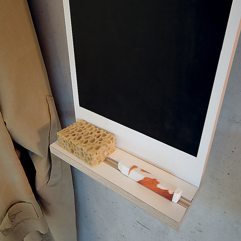 A wall-mounted Moormann Rechenbeispiel features a chalkboard with a wooden shelf below and a nearby storage hook rail. On the shelf, find a sea sponge, chalk on an orange-white striped paper towel, and part of a tan coat in view.