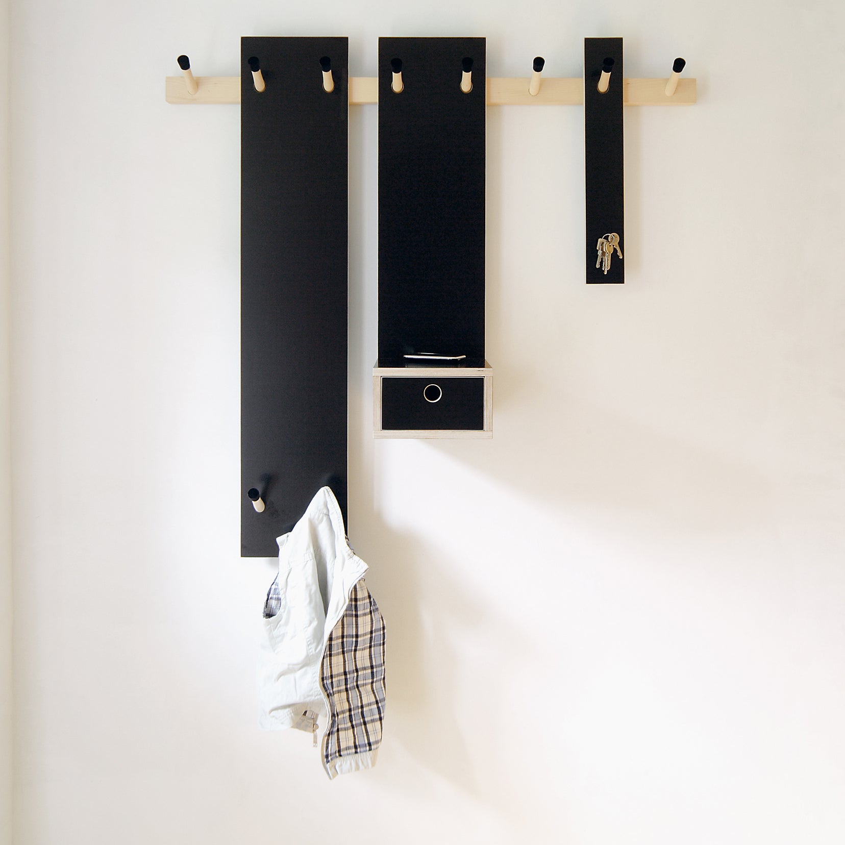 Moormanns Rechenbeispiel is a wall-mounted coat rack with three black panels, wooden pegs, a hook rail, and a small drawer. A plaid shirt hangs over one peg, its minimalist design standing out against the white wall.