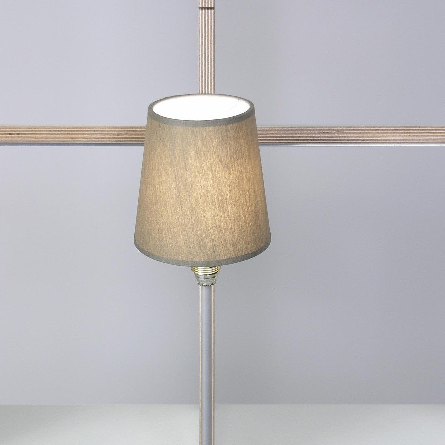 The Rosi Lamp by Moormann, featuring a beige cylindrical shade, lights up the light gray background. Its metal base and pole pair beautifully with the Nils Holger Moormann Shelf, while a wooden crossbar adds artistic flair in the backdrop.