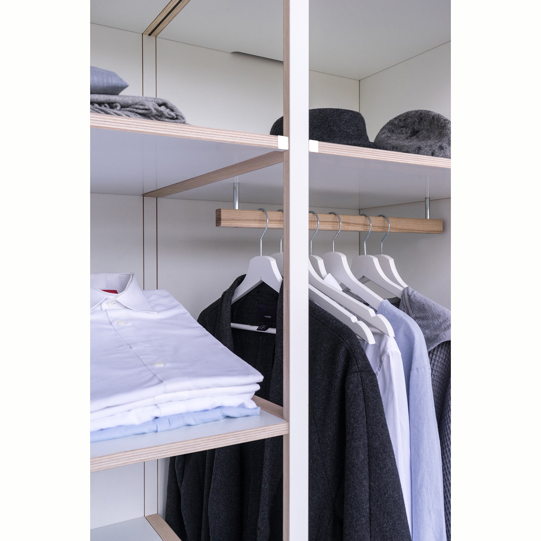 The open wardrobe from Moormanns Schrankone features wooden shelves for folded sweaters and hats on the left, and a modular system with white hangers for shirts, a coat, and a blazer on the right. Its minimalist interior of light-colored wood reflects Schrankones sleek design ethos.