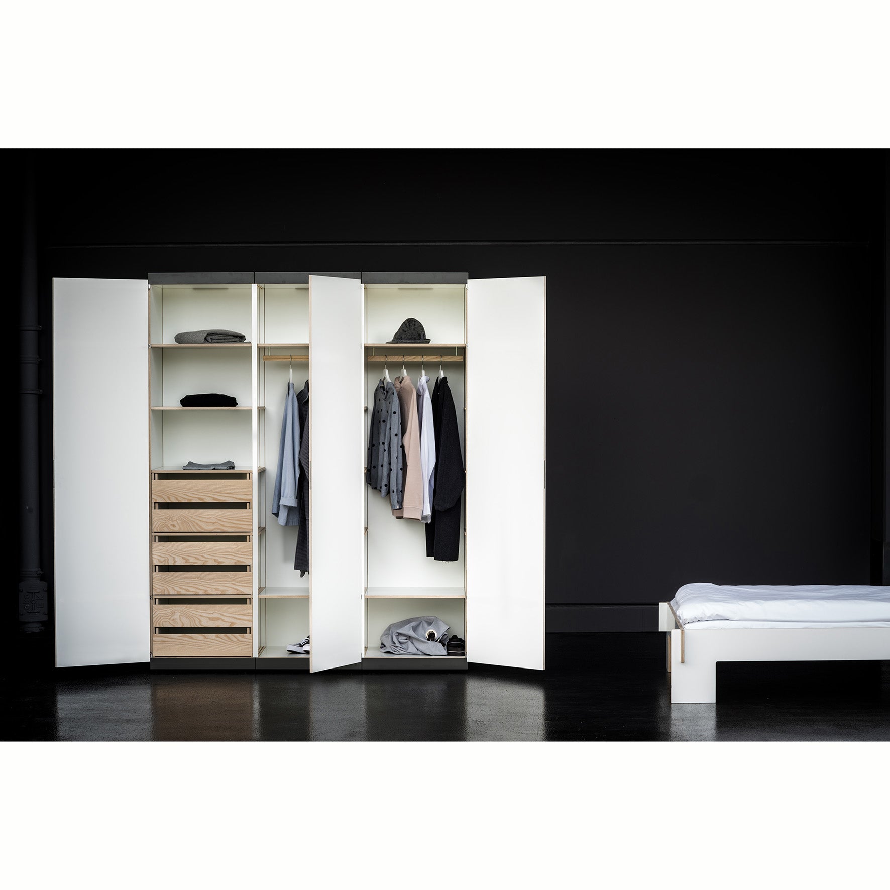 Moormanns Schrankone wardrobe is open against a dark wall, revealing shelves with folded clothes and drawers on the left, hanging shirts and jackets in the center, and shoes at the bottom. A bed with white bedding is partially visible on the right.