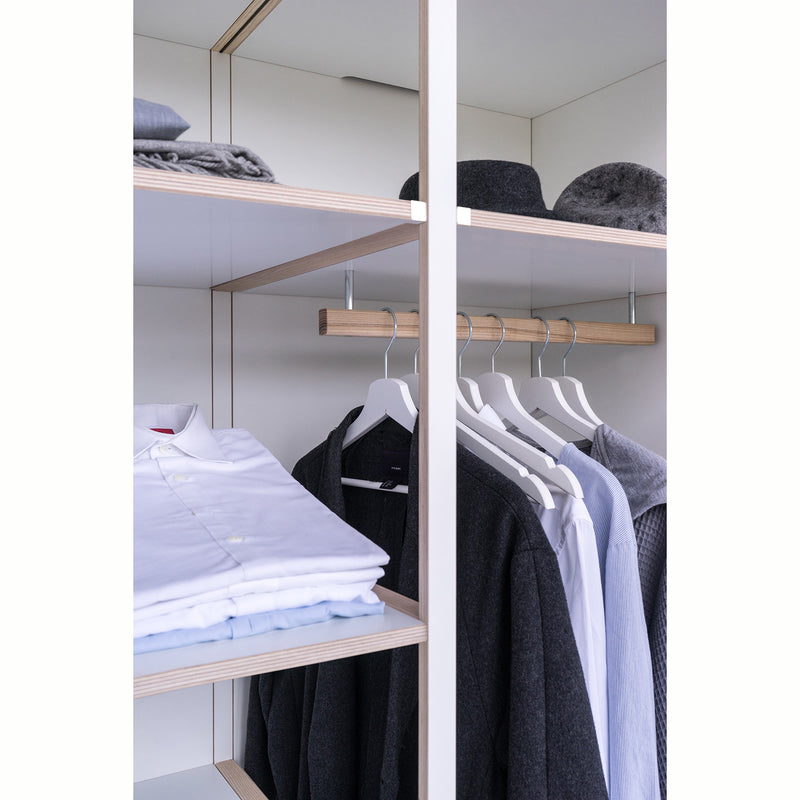 The open wardrobe from Moormanns Schrankone features wooden shelves for folded sweaters and hats on the left, and a modular system with white hangers for shirts, a coat, and a blazer on the right. Its minimalist interior of light-colored wood reflects Schrankones sleek design ethos.