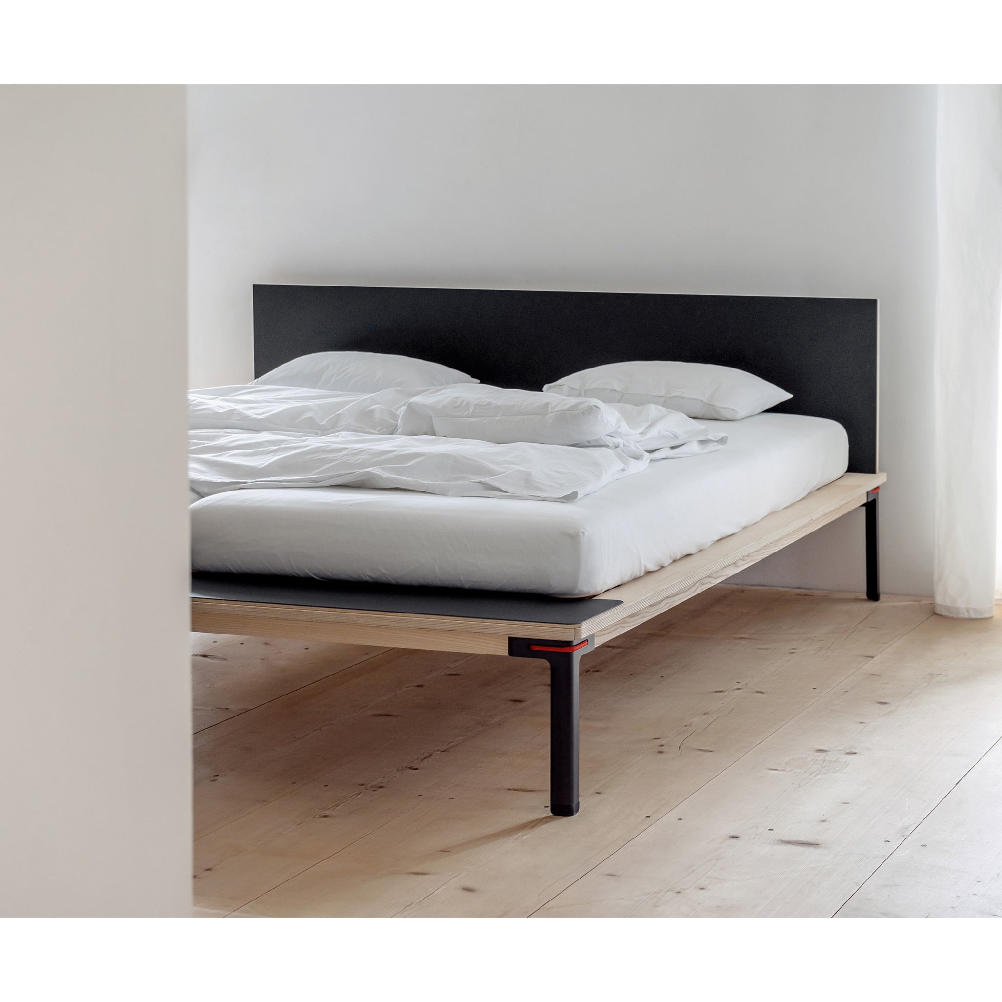 The minimalist bedroom highlights a Moormann Seiltänzer bed with delicate steel legs and a wooden platform, complemented by white bedding and two pillows. It sits on wooden flooring with a dark headboard against a white wall, basking in natural light for a serene atmosphere.