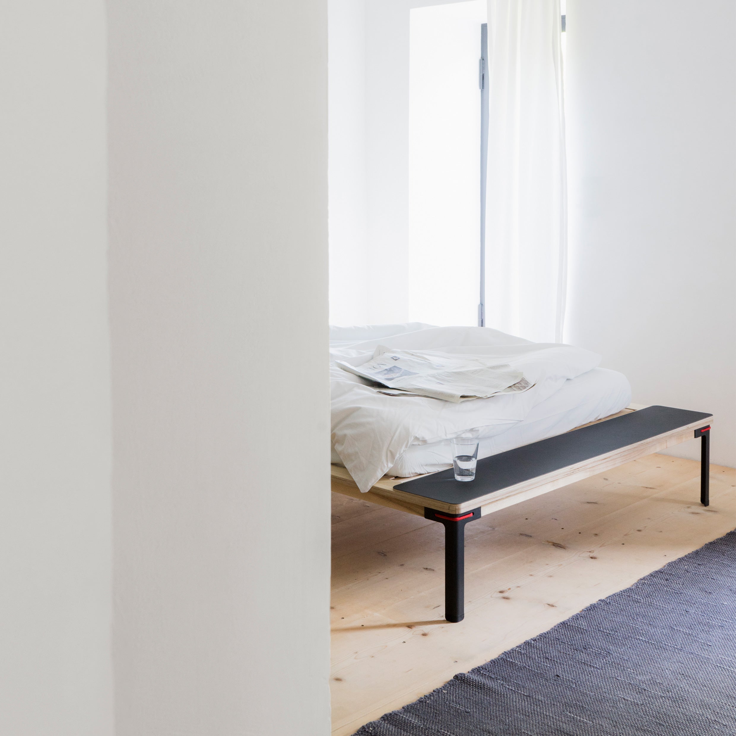 A minimalist bedroom features Moormanns Seiltänzer bed with sleek filigree steel legs, accompanied by white bedding. A glass of water sits on a black bench at the foot. Sheer curtains filter natural light onto the light wooden floor, creating an airy vibe.