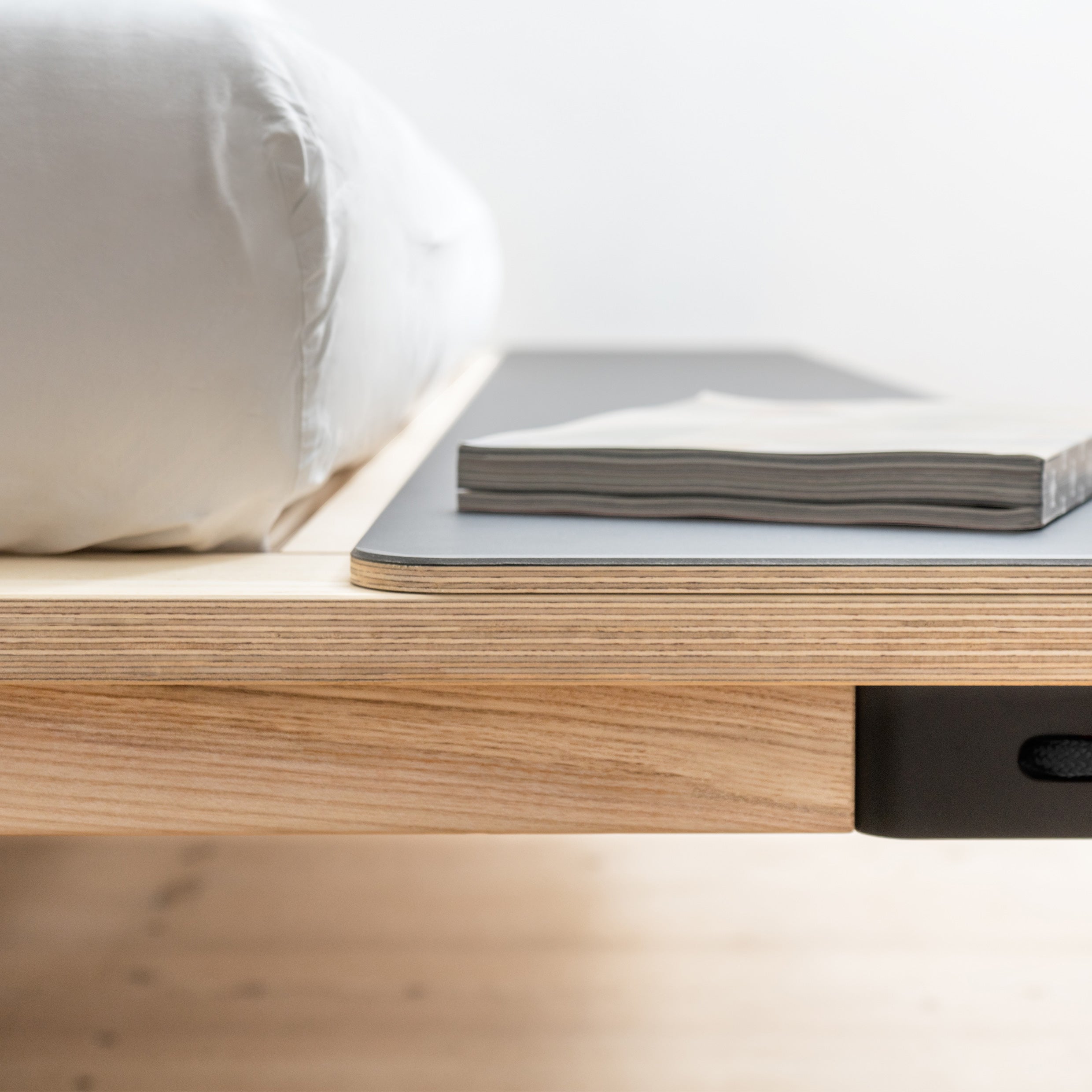 Close-up of Moormanns Seiltänzer Bed, showcasing its minimalist design with filigree steel legs. A book sits on the side shelf of its floating frame. White bedding covers the mattress, and the softly blurred background enhances its elegant look.