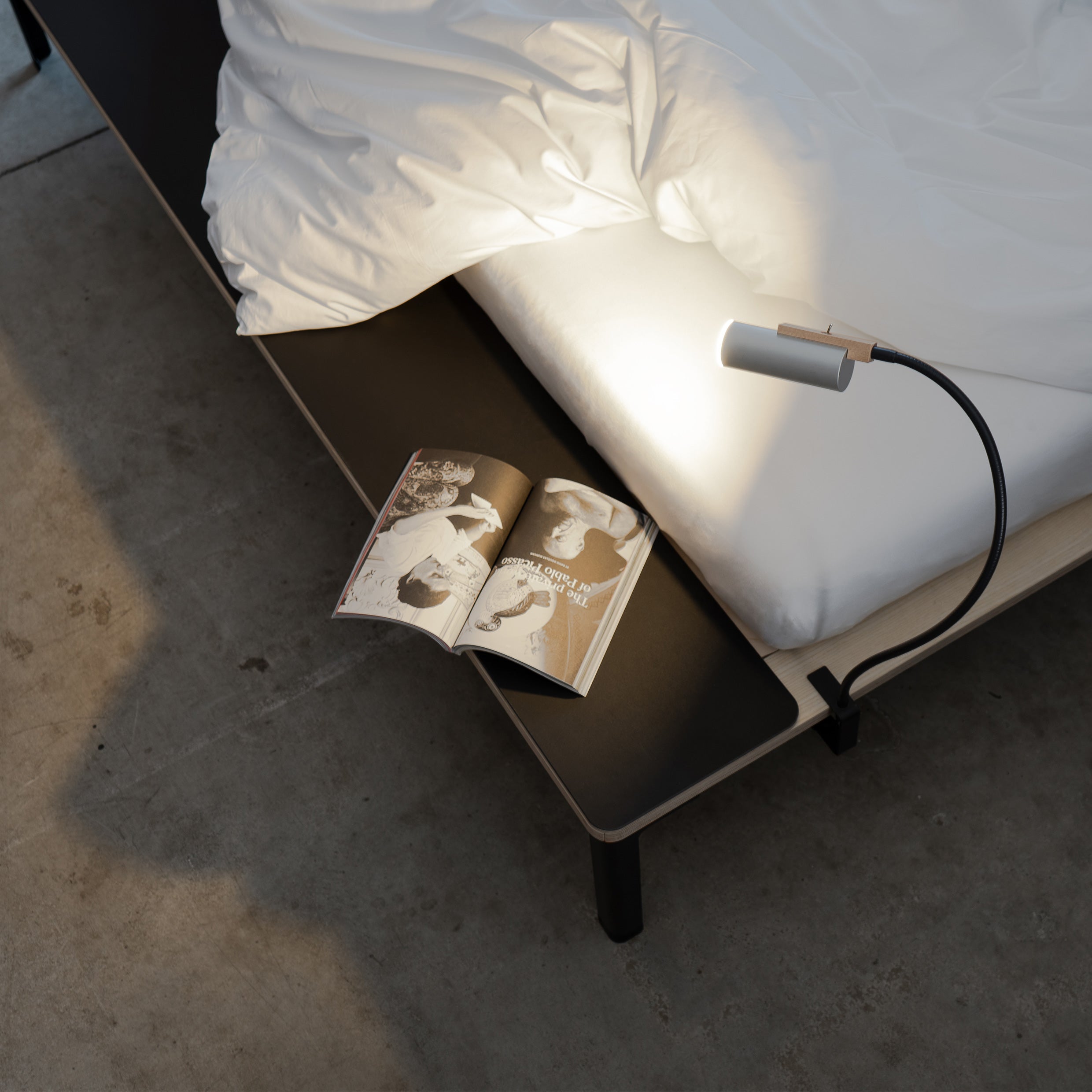 A Moormann Seiltänzer Bed with white sheets rests on a wooden platform with filigree steel legs. An open magazine under a flexible reading lamp, and sunlight streaming onto the concrete floor creates an airy atmosphere around the floating surface.