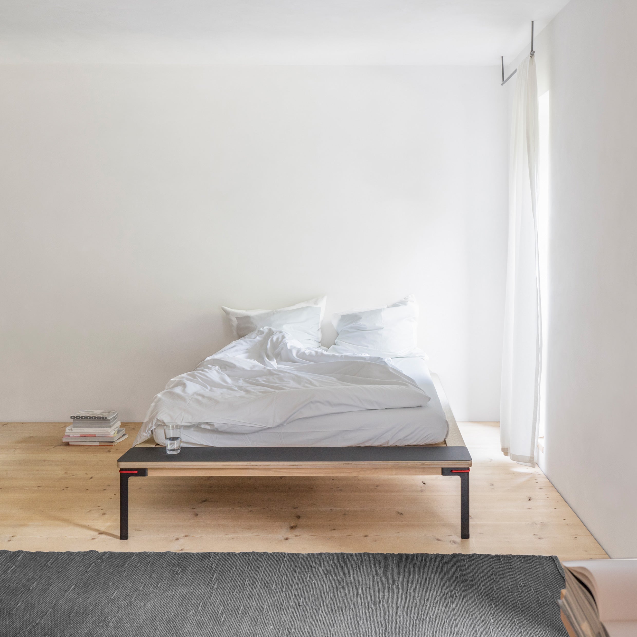 In the minimalist bedroom, a Moormann Seiltänzer Bed with sleek filigree steel legs and a modern low frame takes center stage. The floating surface is adorned with white bedding and pillows, while books and a glass of water sit on the floor. Natural light filters through onto a dark rug in front.
