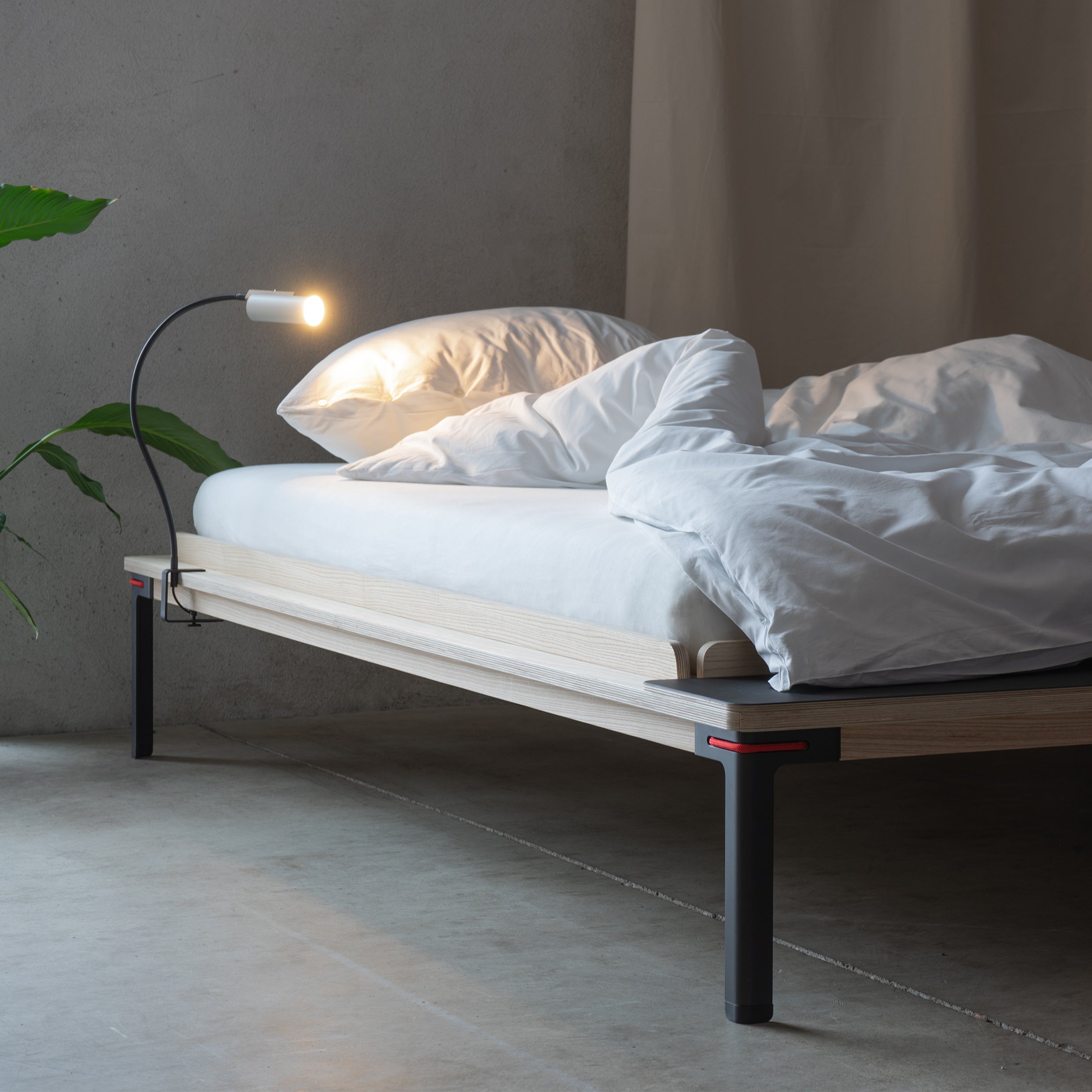 The minimalist bedroom showcases Moormanns Seiltänzer Bed with white bedding and a thin mattress. A small lamp lights the bed on filigree steel legs, while a green plant adorns the softly lit room with concrete flooring and a neutral backdrop.