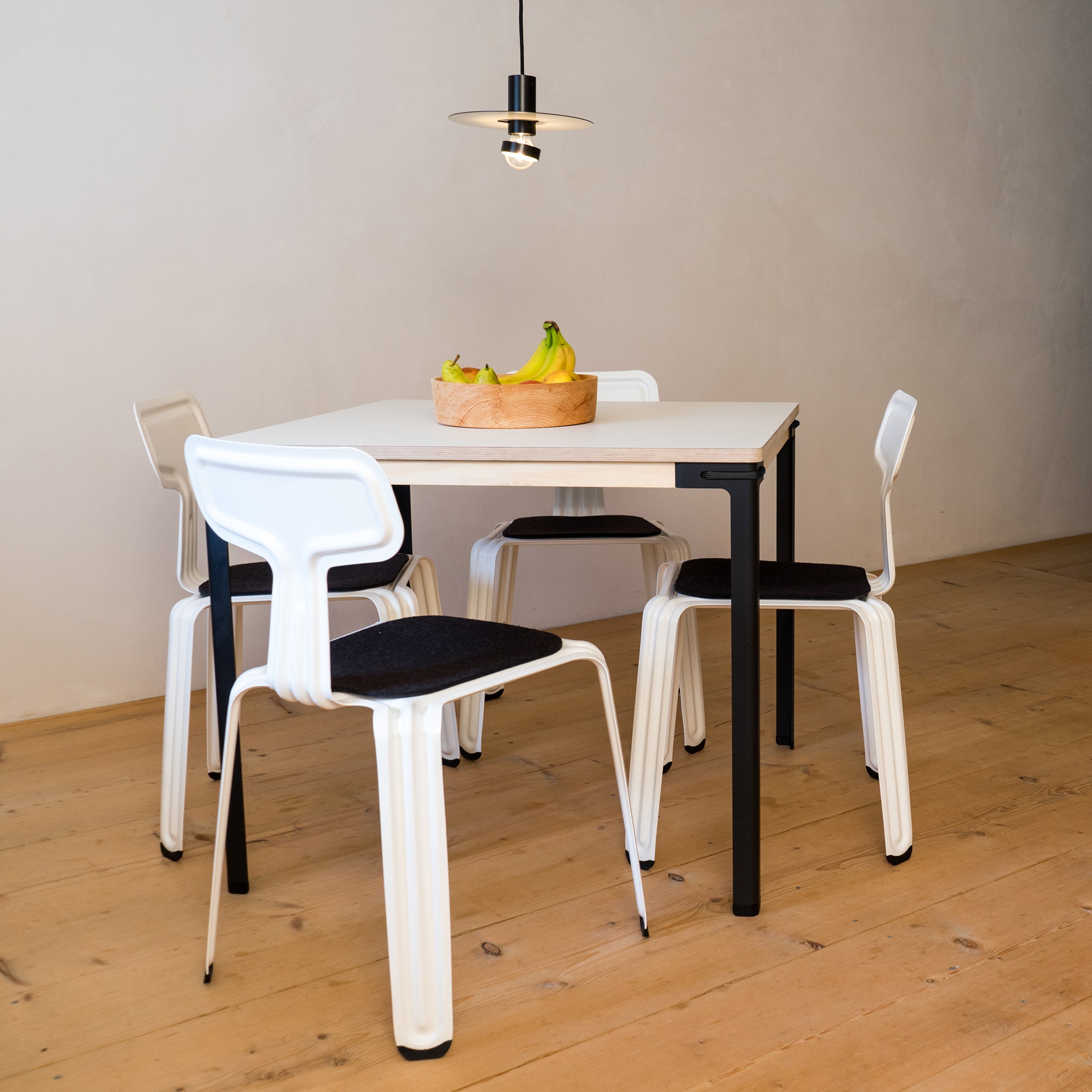 In a minimalist dining area, a Moormann Seiltänzer Table with steel legs is surrounded by four white chairs with black seats. A wooden bowl of bananas sits on top, while a black pendant light floats above. The light wood floor and plain wall complete the airy ambience.