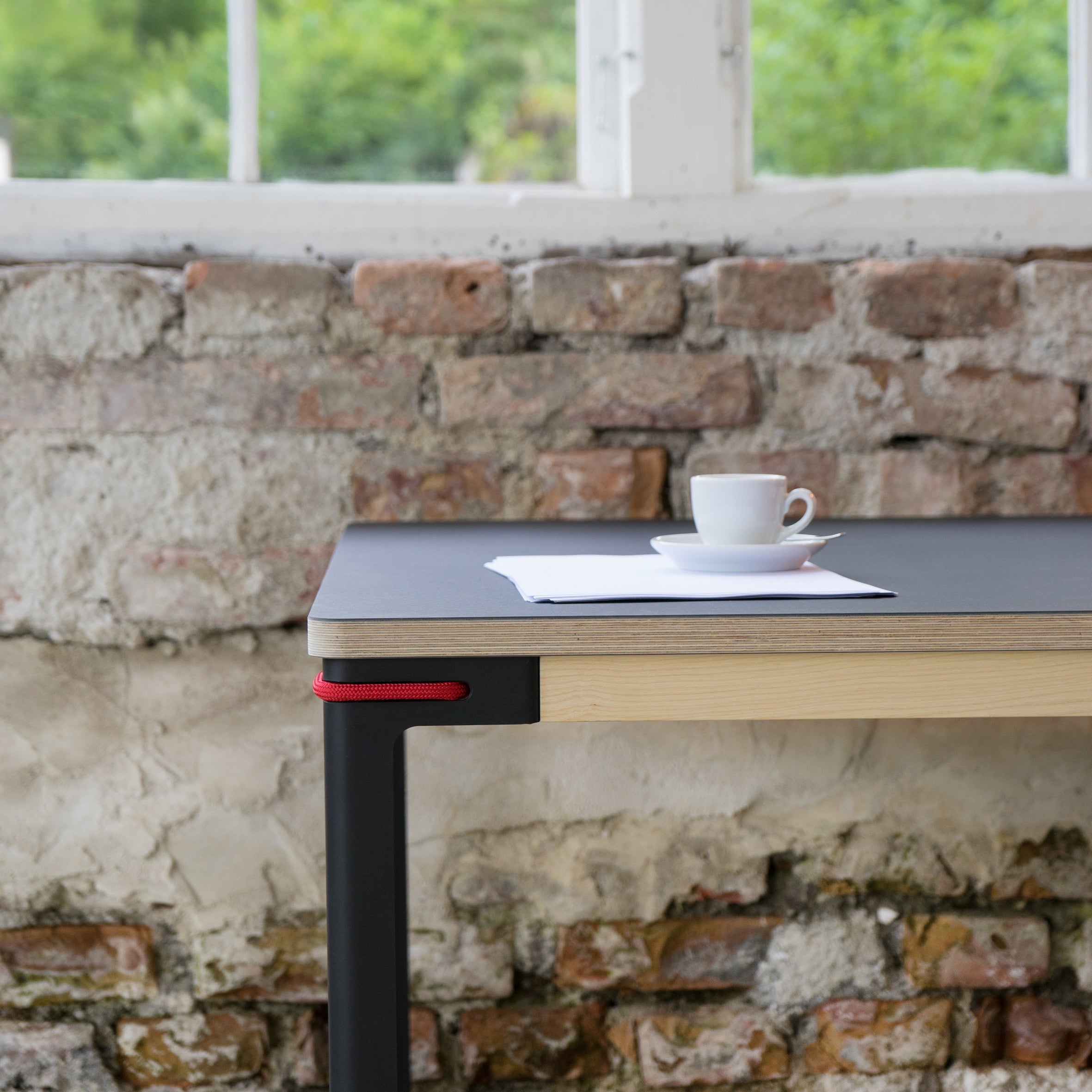 A Moormann Seiltänzer Table - SALE with steel legs and black top stands by a textured brick wall. A white cup, saucer, and paper rest on it while natural light from a large window above reveals greenery outside. The table features a red accent on the corner.