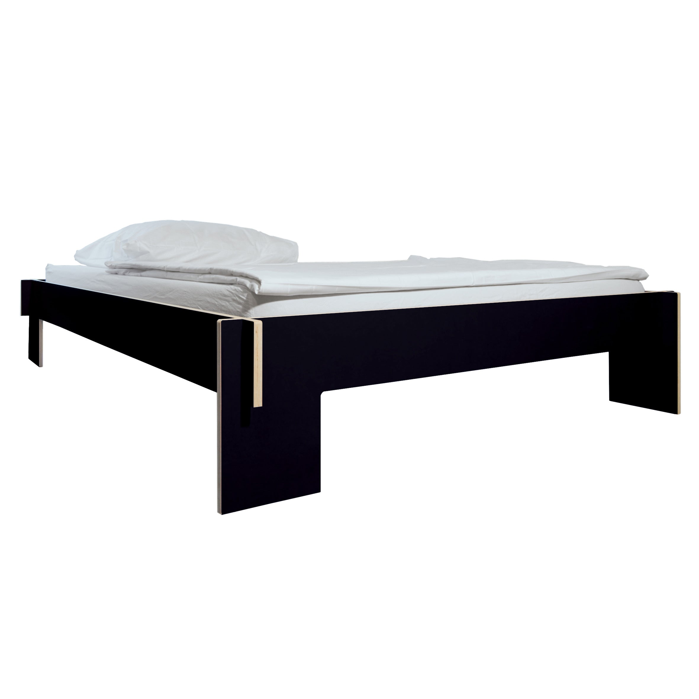 The Siebenschläfer by Moormann is a minimalist black platform bed made from birch plywood. It features a simple, rectangular frameless design with white pillow and sheet, set against a white background. The flat-packed bed lacks a headboard and footboard.