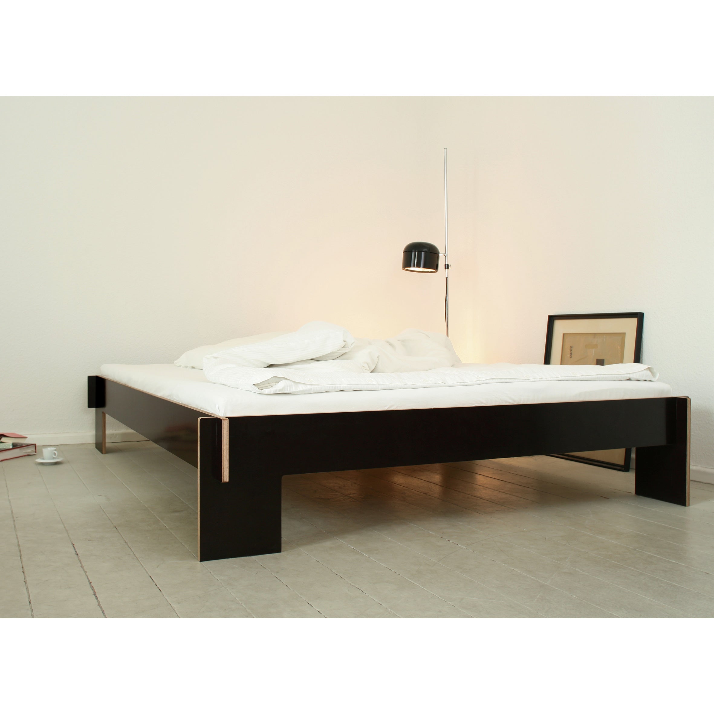 A minimalist bedroom showcases a sleek, flat-packed Siebenschläfer bed by Moormann, crafted from birch plywood and requiring no tools for assembly. The low black platform contrasts elegantly with the white bedding. Nearby, a modern black floor lamp stands next to a stylishly leaned framed picture.