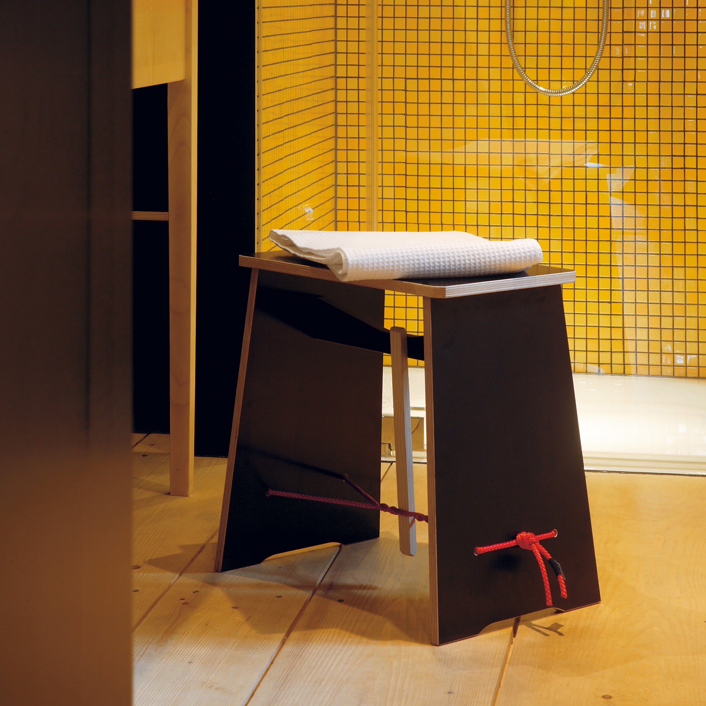 A modern black Moormann stool with a slanted design stands on a wooden floor. A red cord ties around one leg, and a white towel rests on top. The yellow square tiled background and part of a shower setup evoke mornings reminiscent of Strammer Max.