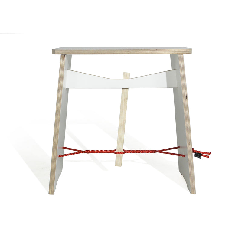 The Strammer Max by Moormann is a minimalist wooden stool made from birch plywood with a flat rectangular seat. Designed by Max Frommeld, it features two parallel legs connected by a red rope and a white horizontal bar, embodying simplicity and modernity.