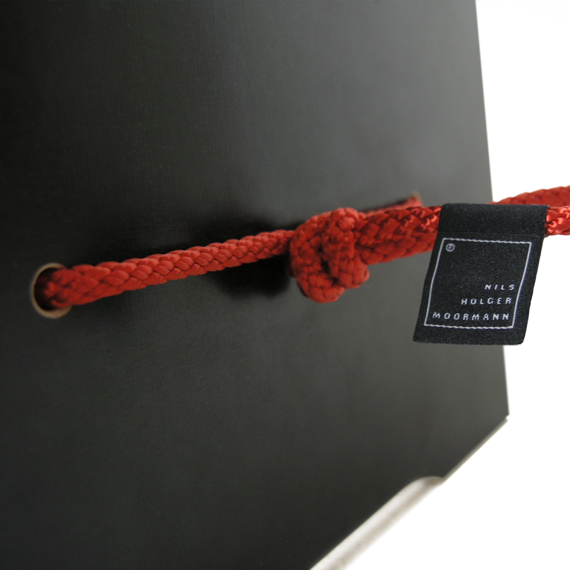A close-up of a red rope knotted through two grommets on a black surface featuring a small black tag reading Nils Holger Moormann. This creative design evokes the innovative Strammer Max by Moormann, renowned for its sleek plywood panels.
