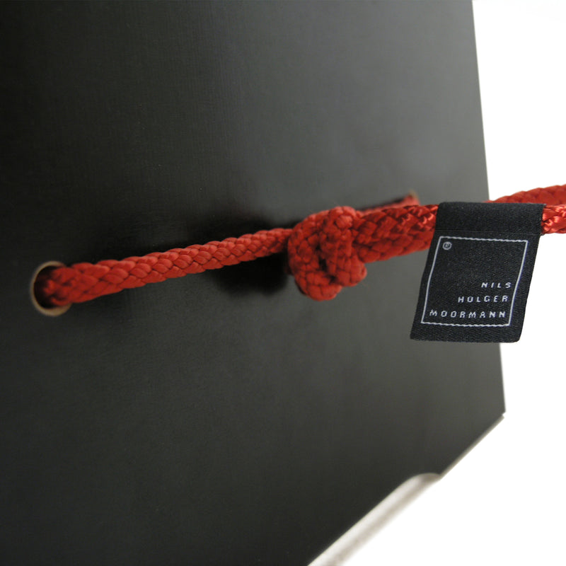A close-up of a red rope knotted through two grommets on a black surface featuring a small black tag reading Nils Holger Moormann. This creative design evokes the innovative Strammer Max by Moormann, renowned for its sleek plywood panels.