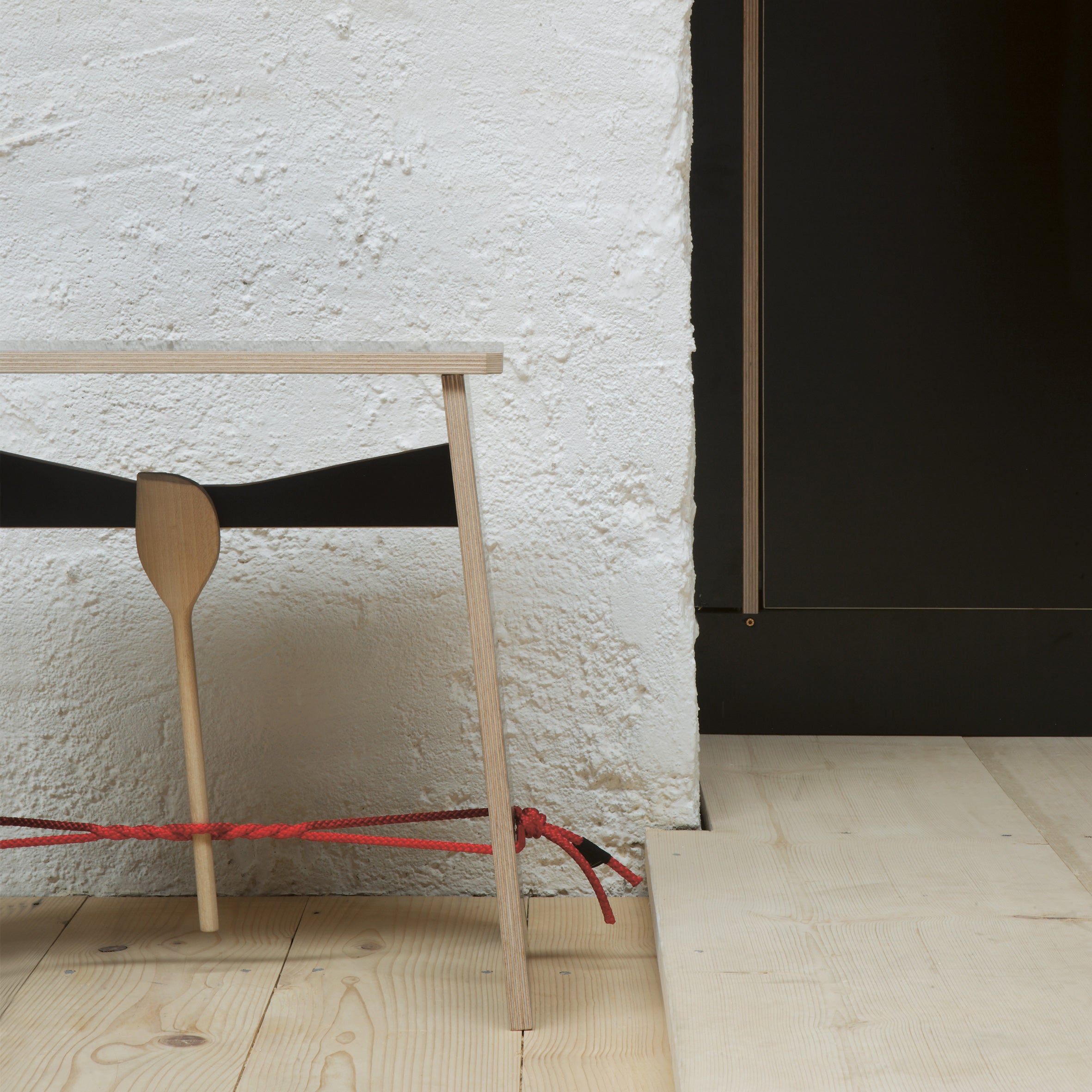 A Moormann Strammer Max minimalist wooden bench stands against a white textured wall, accented by a black strap and wooden paddle. A red rope creatively ties across its legs, complementing the light plywood floor. A dark door is partially visible to the right.