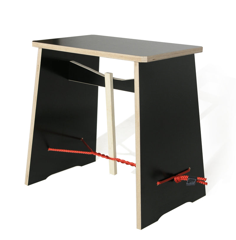The Strammer Max by Moormann is a creatively designed stool with a minimalist charm, featuring a black seat and sides made of plywood. A red decorative rope weaves through the base, adding flair, while a wooden crossbar ensures stability.
