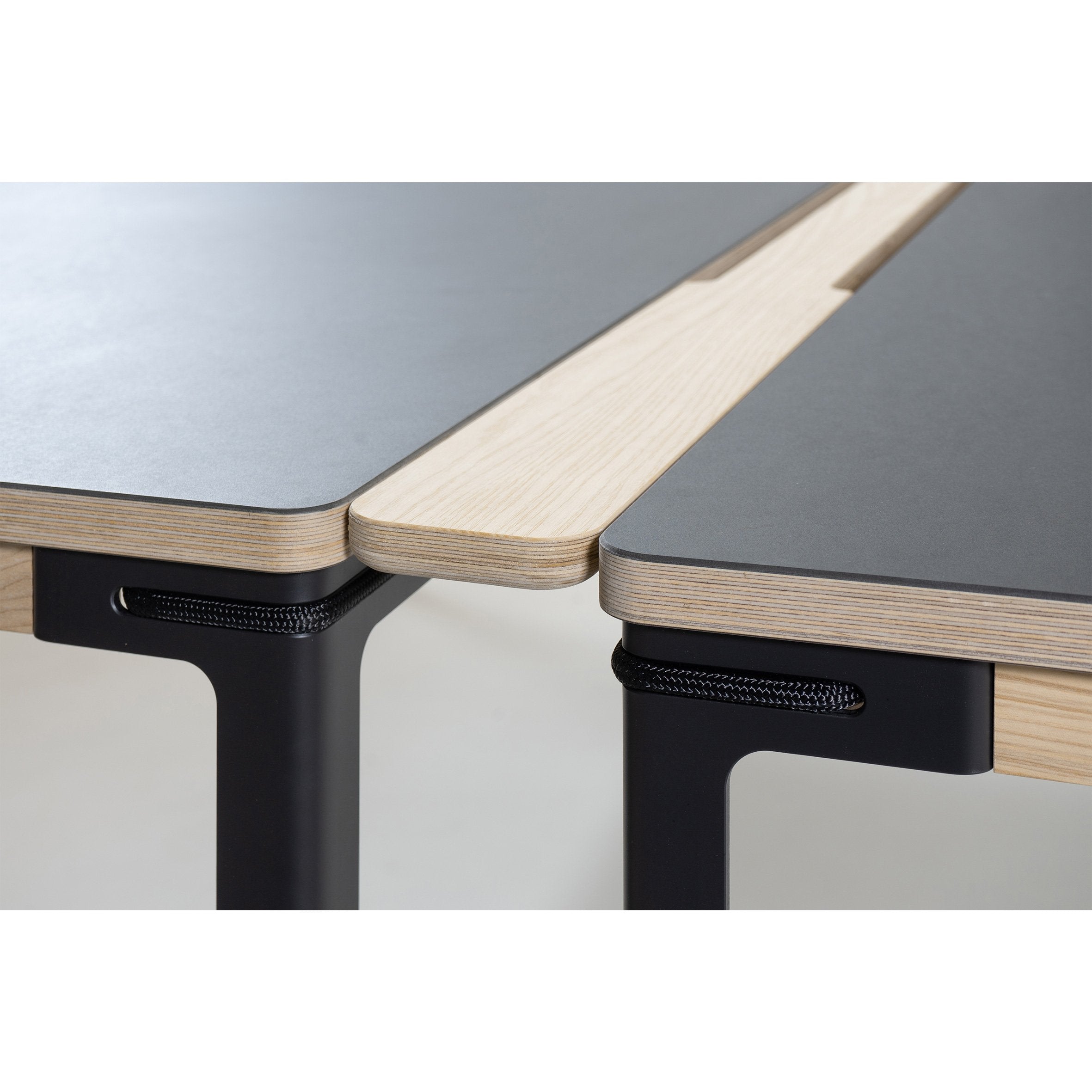 The image showcases a modular office desk system, featuring a light wood finish and black surfaces. It highlights the seamless connection of two desk sections, similar to Moormanns Table Connector design, with dark metal supports and integrated cable passage management for a modern look.