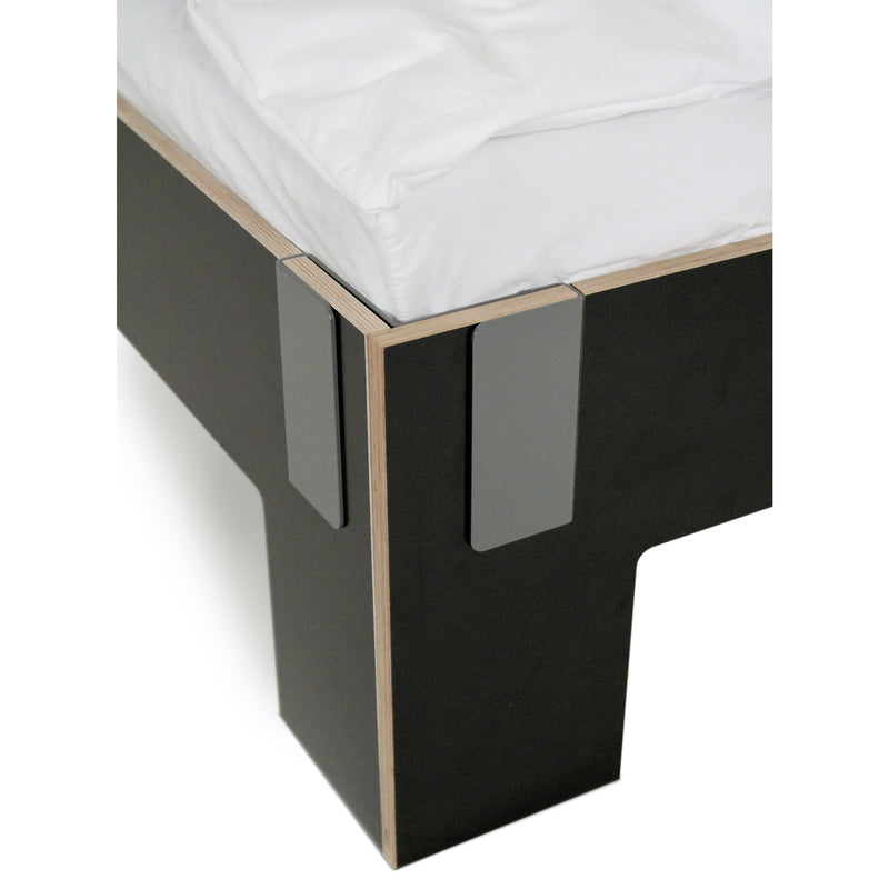 The Tagedieb bed by Moormann features a sleek black frame with exposed plywood edges. Its corners boast a gray metallic bracket design, while a white mattress enhances the minimalist charm of this contemporary piece.