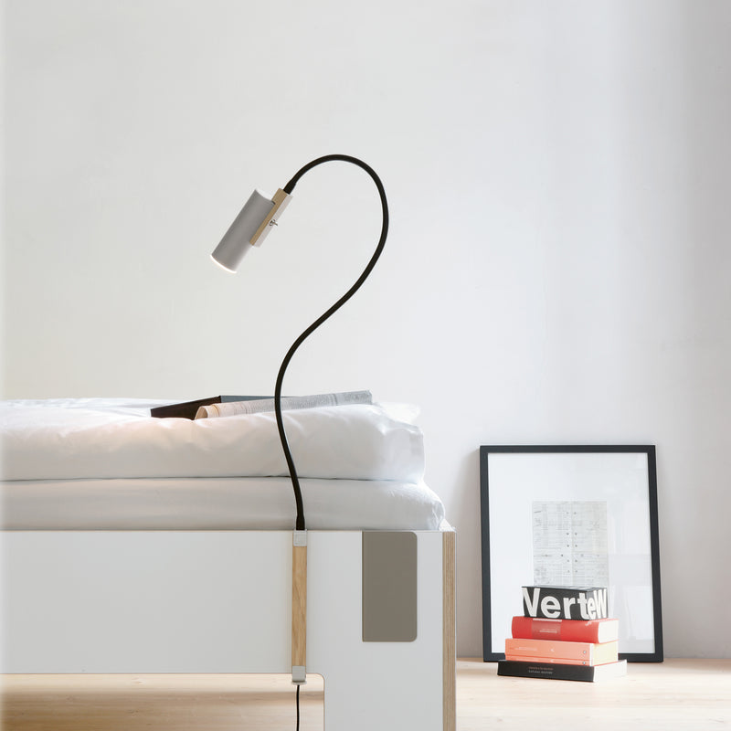 A minimalist bedroom showcases a Moormann Tagedieb bed with adjustable mattress support. The white bed, topped with fresh bedding, is paired with a flexible black lamp illuminating an open book. Nearby, a black-framed picture and books, including Verte, rest on the wooden floor.