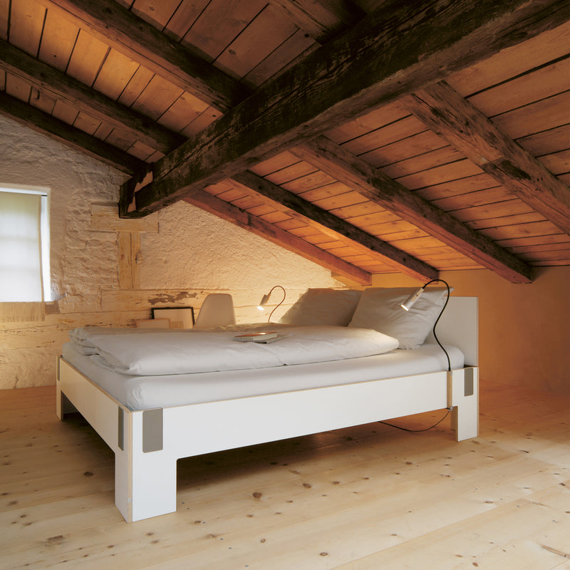 In a cozy attic bedroom, wooden slanted ceiling beams and a light wood floor create warmth. Against a stone wall rests Moormanns Tagedieb bed with its high headboard and gray bedding, softly lit by modern lamps. Natural light streams through a small window, enhancing the rooms serene ambiance.
