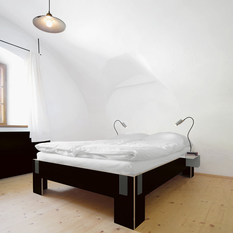 A minimalist bedroom showcases a Moormann Tagedieb bed with adjustable mattress support, a black frame, and white bedding. Two reading lamps flank the high headboard. The room features wooden floors, a white arched ceiling, and light curtains over the window.