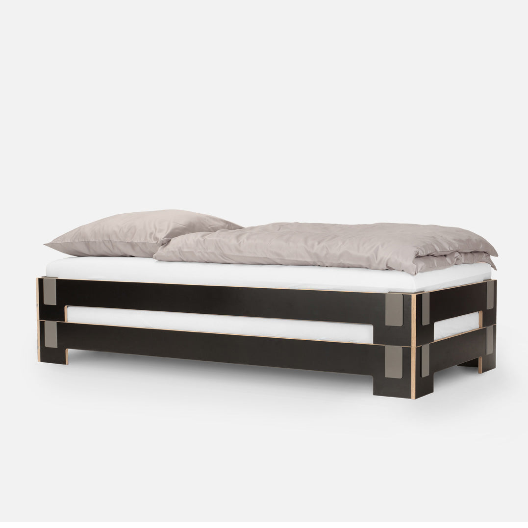 Experience the sleek design of Moormanns Tagedieb Stackable bed, featuring a minimalist black frame with beige accents. It includes a white mattress, two gray pillows, and a gray duvet. Its flexible headboard offers versatility against a plain white backdrop.